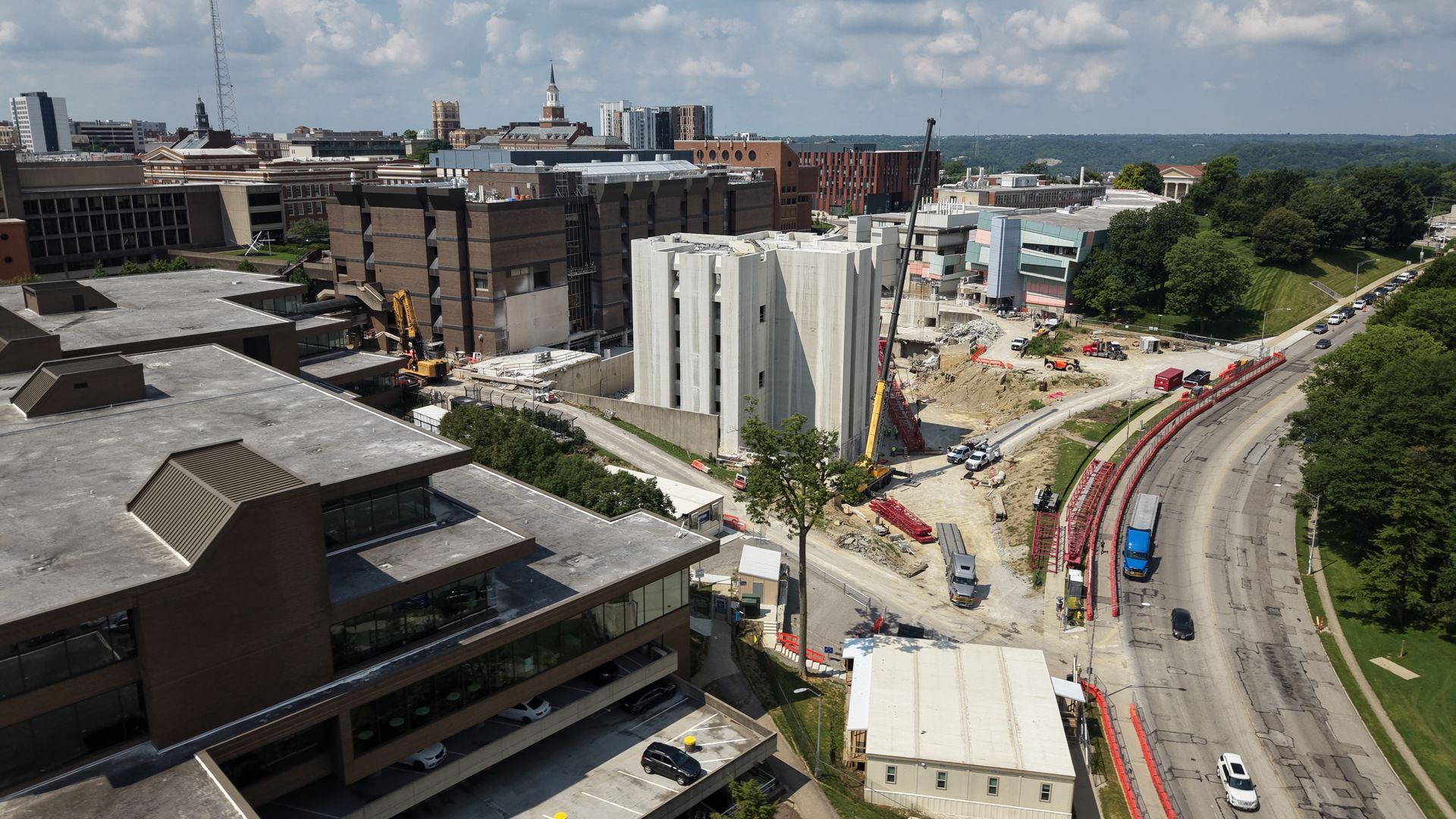A wide aerial view of a city: modern brick buildings, a large construction site with cranes, a curved road lined with red barriers, green trees on the right, and a blue sky with fluffy clouds.