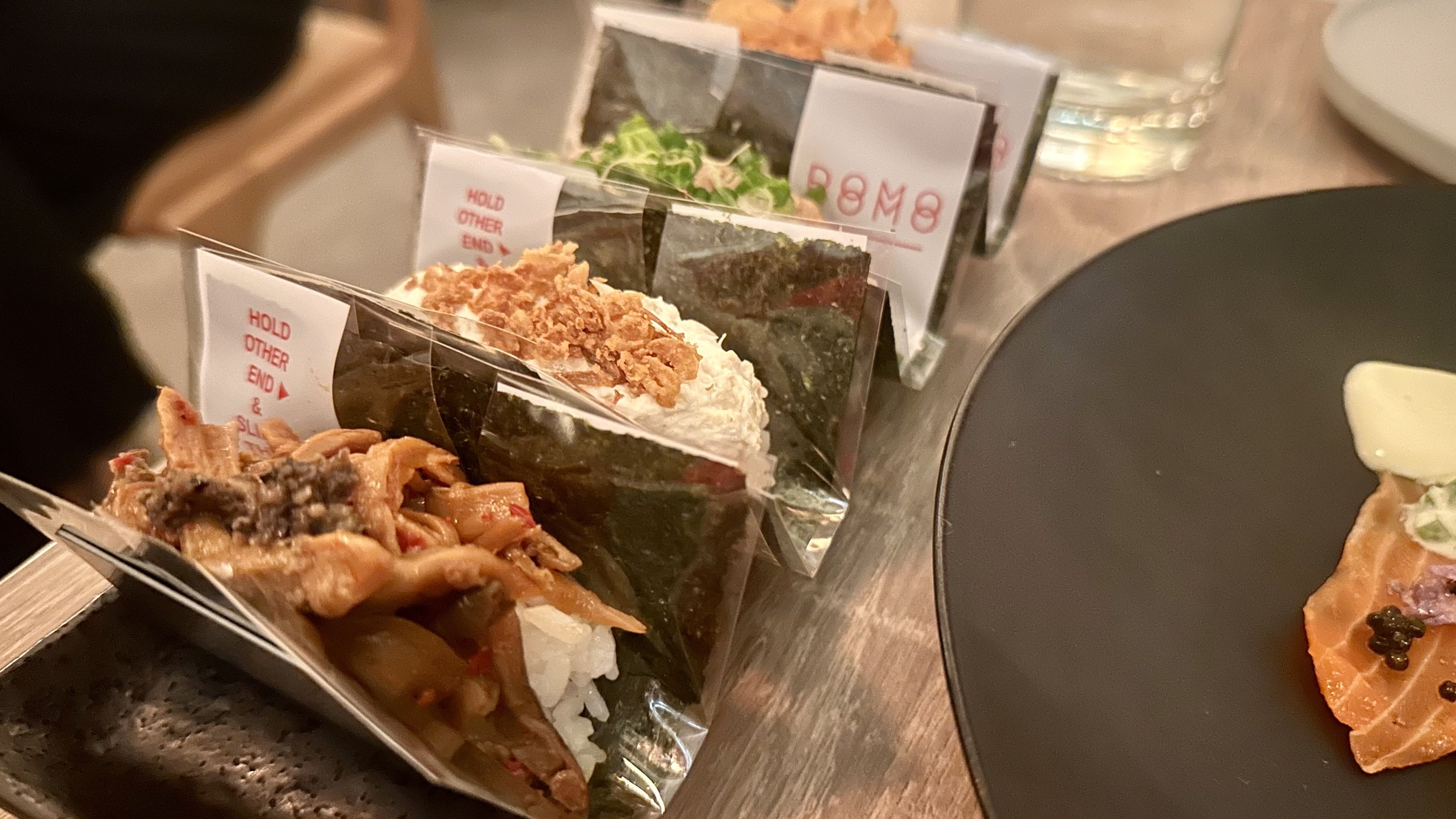 Row of wrapped sushi cones on a wooden table, each in plastic sleeves. A dark plate on the right holds salmon sashimi and greens under warm lighting.
