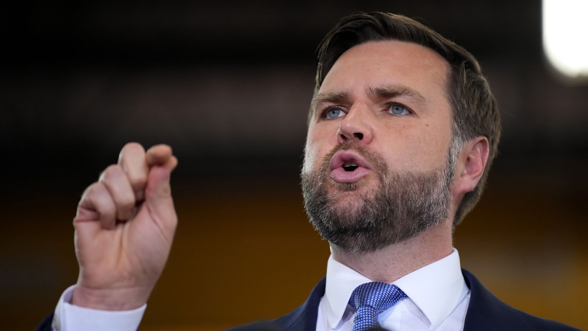 JD Vance holds his hand up as he speaks, wearing a dark suit and a white shirt.