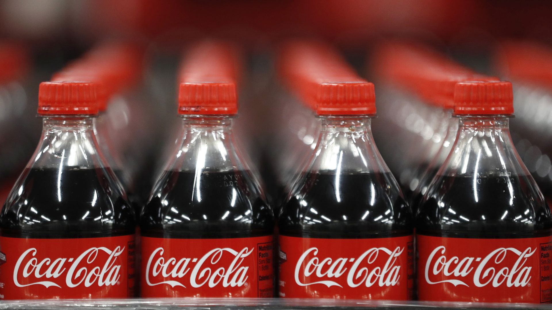 Rows of Coca-Cola plastic bottles with red caps and labels, filled with dark soda, displayed on a store shelf in a close-up view.