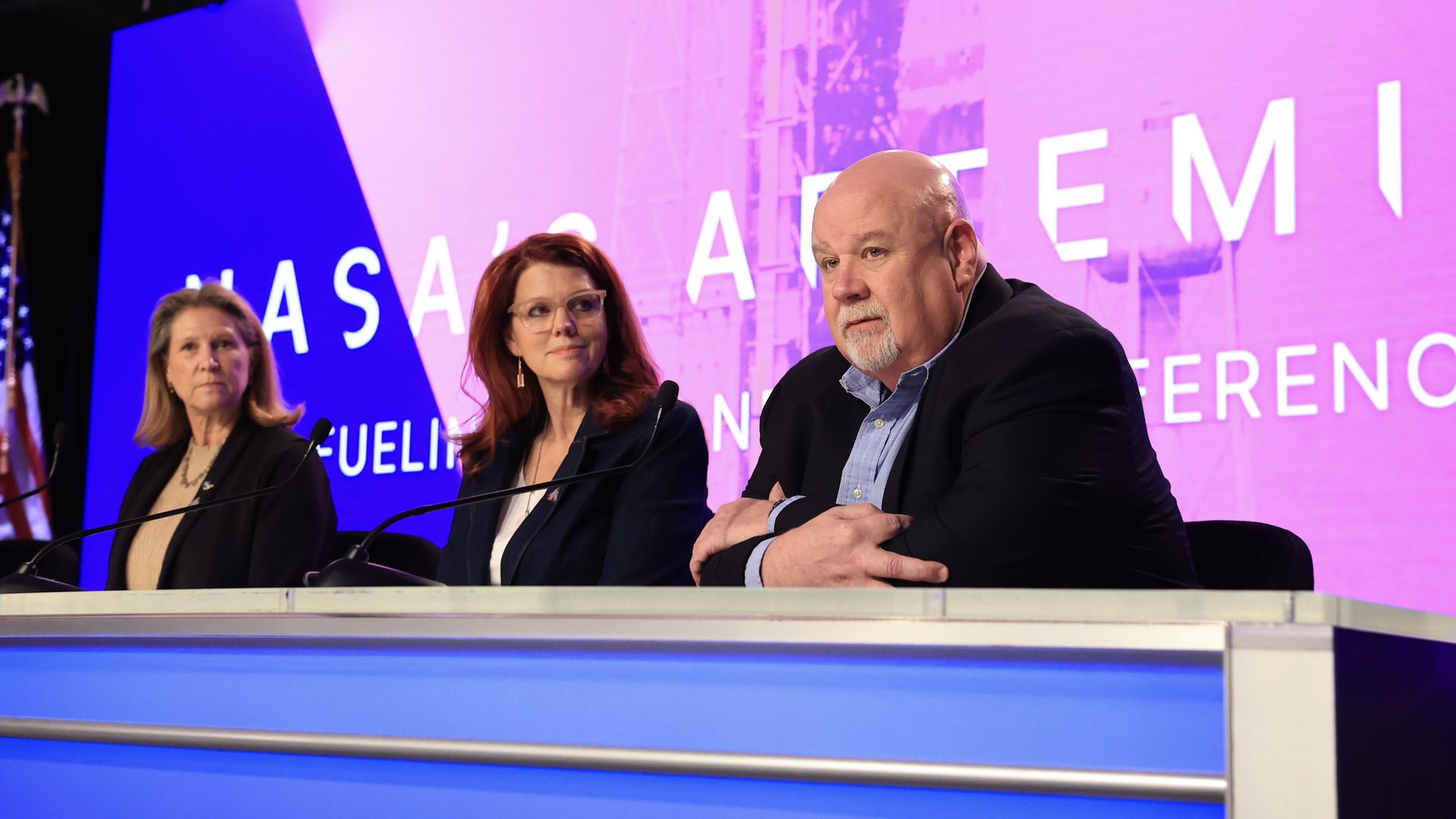 Three people sitting behind a blue desk with microphones at a NASA Artemis event with a pink and blue background showing the NASA Artemis logo.