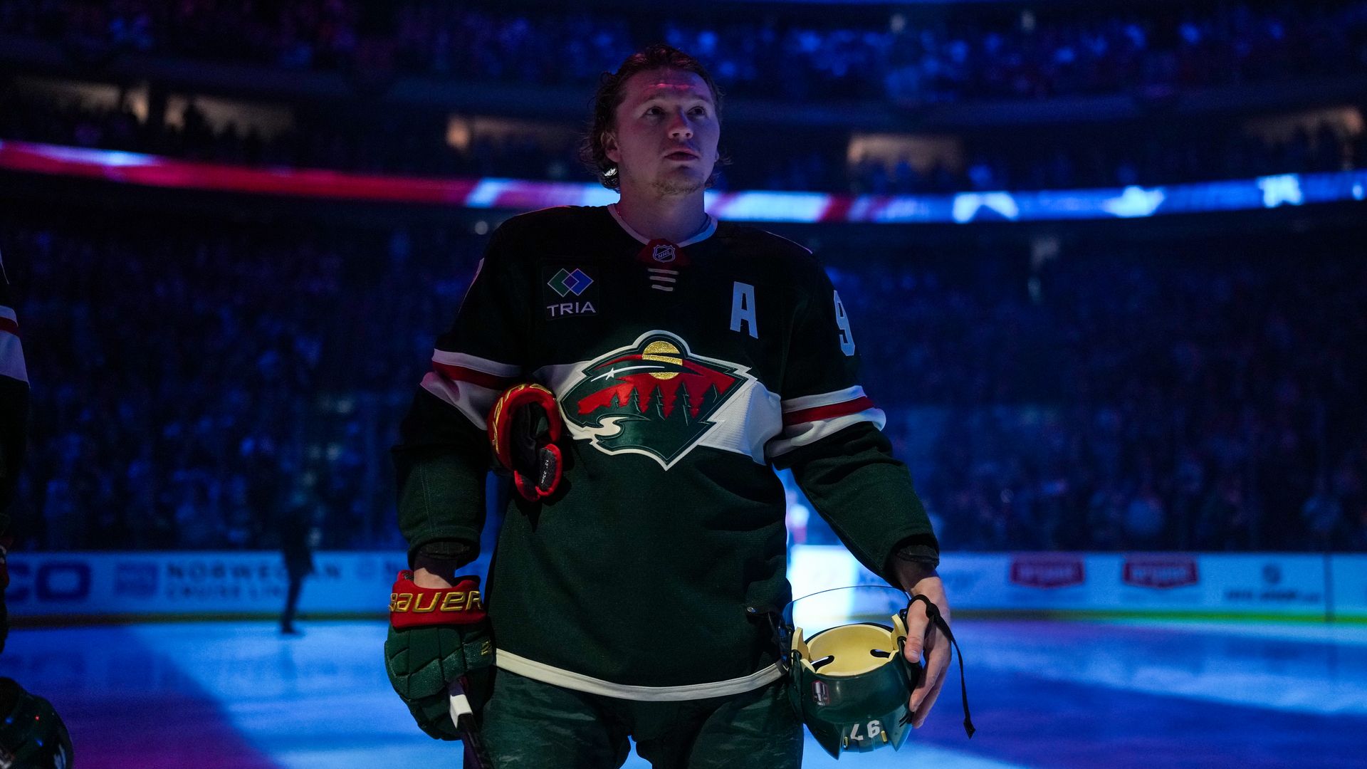 A hockey player in a green Minnesota Wild jersey on the ice in half-light during the US national anthem