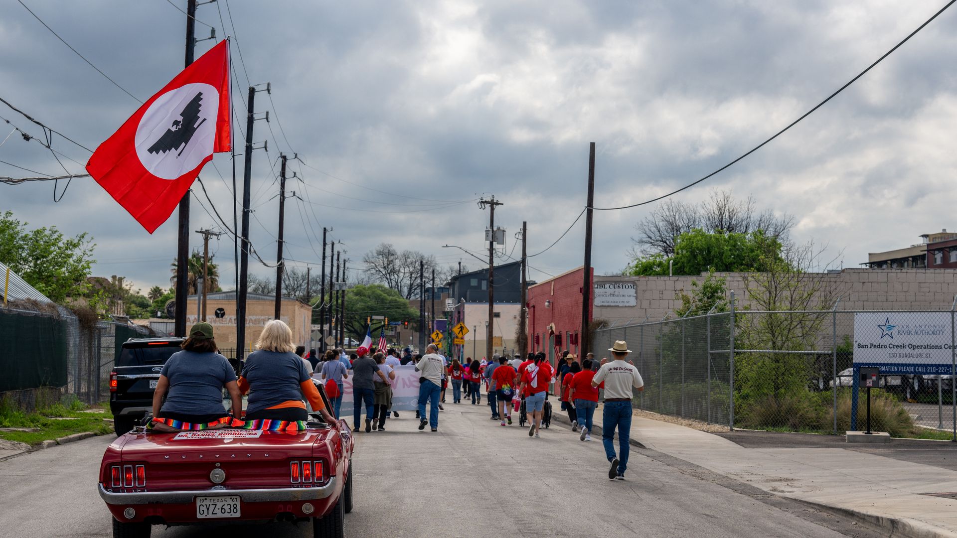 Red vintage Ford Mustang convertible with two riders leads a city street parade; a large red flag with a white circle and black eagle waves as a crowd in red shirts walks down a street under a cloudy sky.