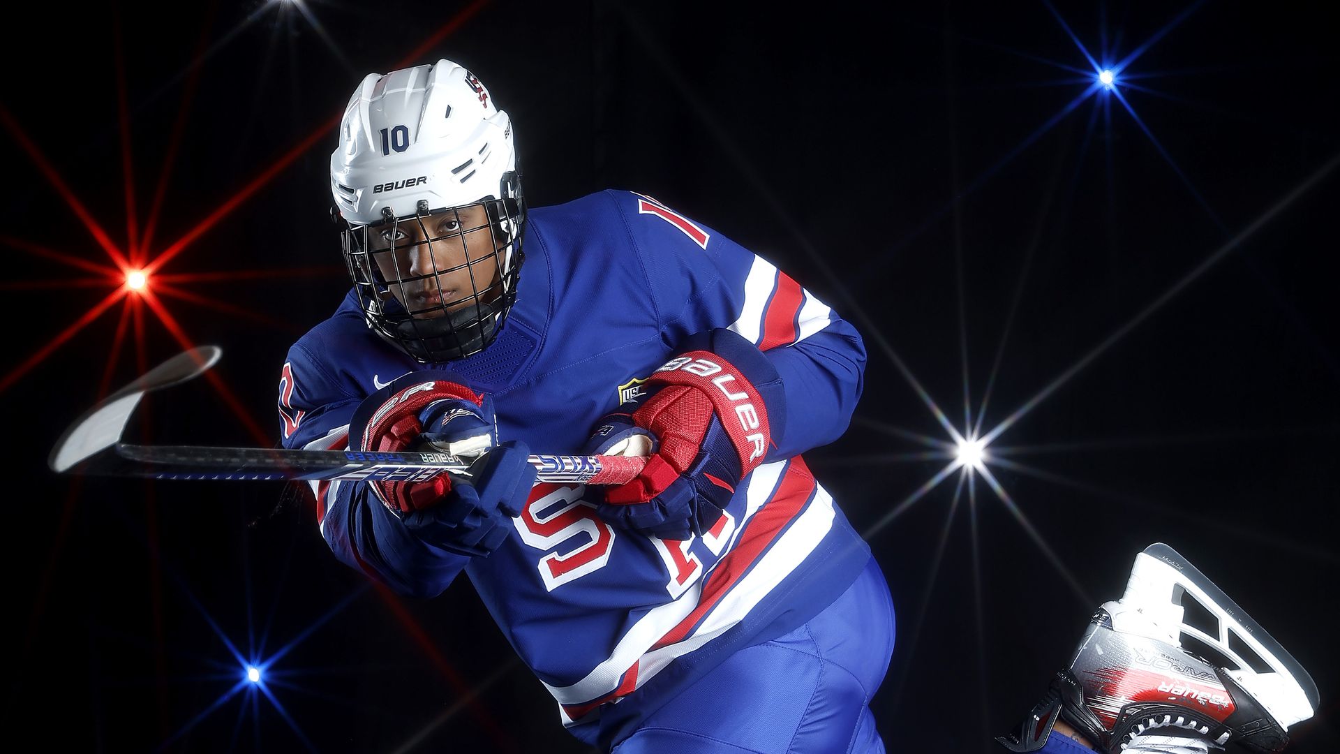 Hockey player in blue, red, and white USA uniform, white helmet with number 10, in mid-action pose holding a stick, against black background with red, white, and blue star-like lights.