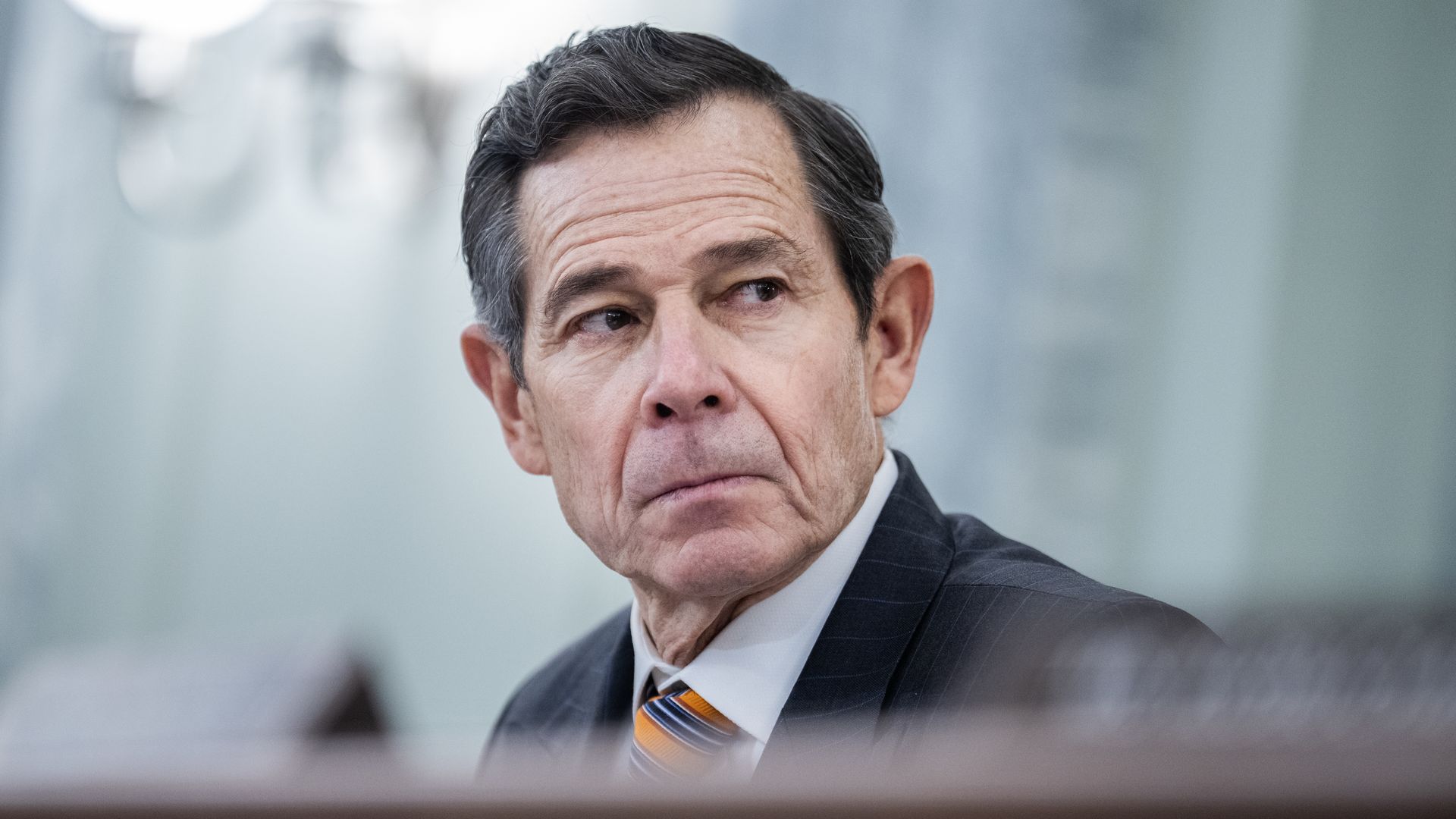 An older man in a dark pinstriped suit, white shirt, and orange-striped tie looks to his left with a serious expression, set against a blurry, light blue background.