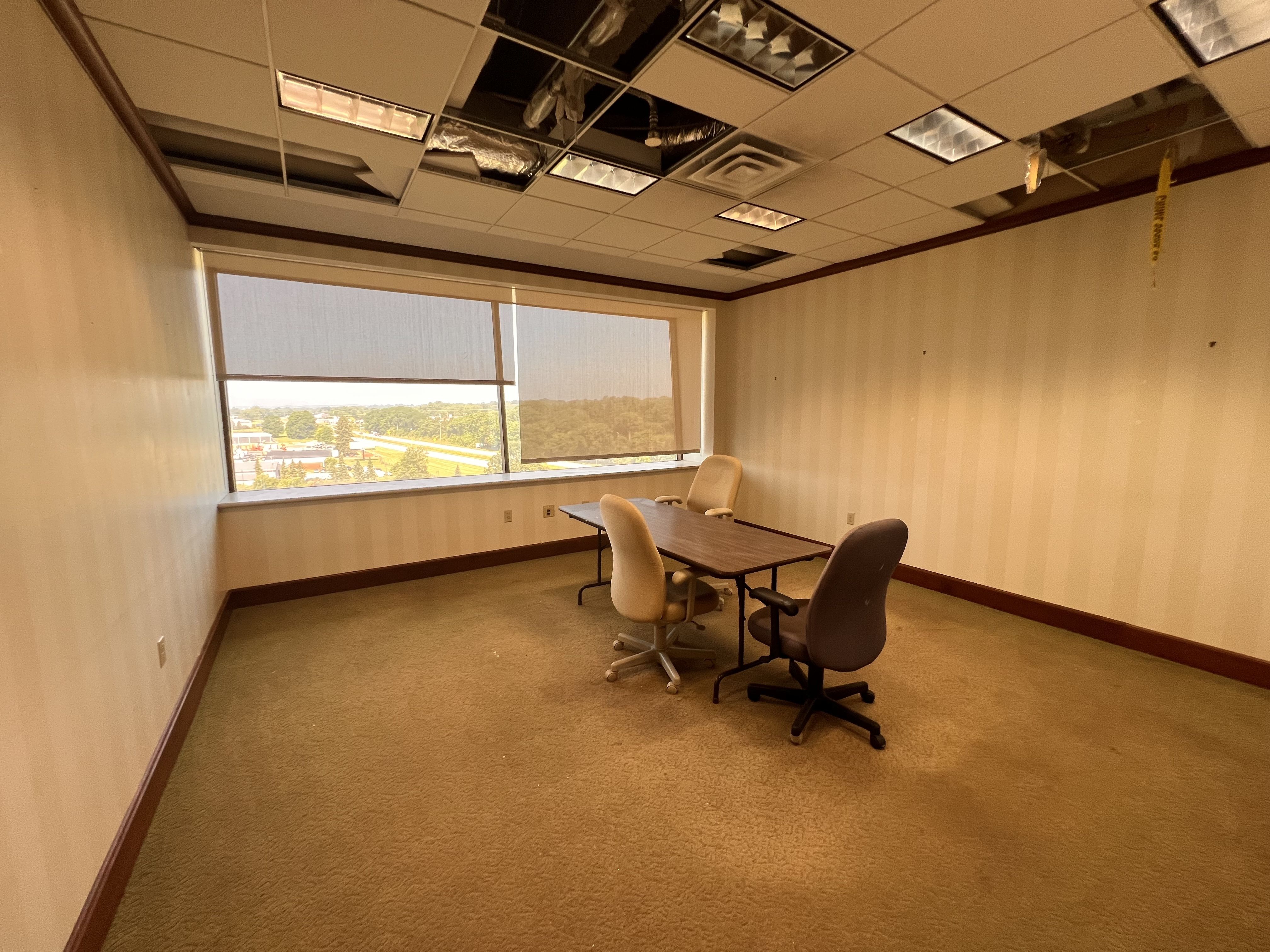Conference room with a damaged drop ceiling, exposing ducts and missing tiles. Beige walls with brown trim, a large window with an outdoor view. Several chairs surround a wooden table; yellow caution tape along the right wall.