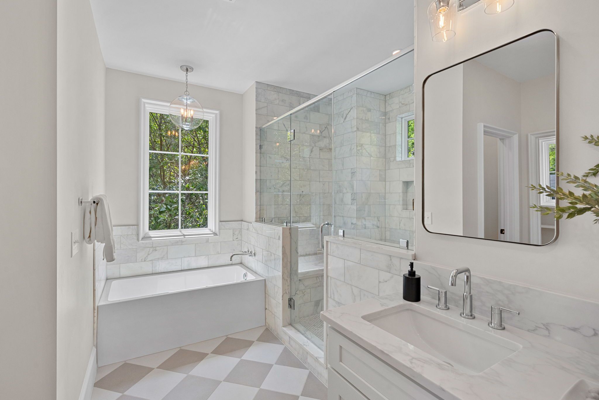 Bright modern bathroom with marble walls, glass shower enclosure, white tub, and a rounded mirror above marble counter. Window shows greenery; pendant lights; grey and white checkered floor.