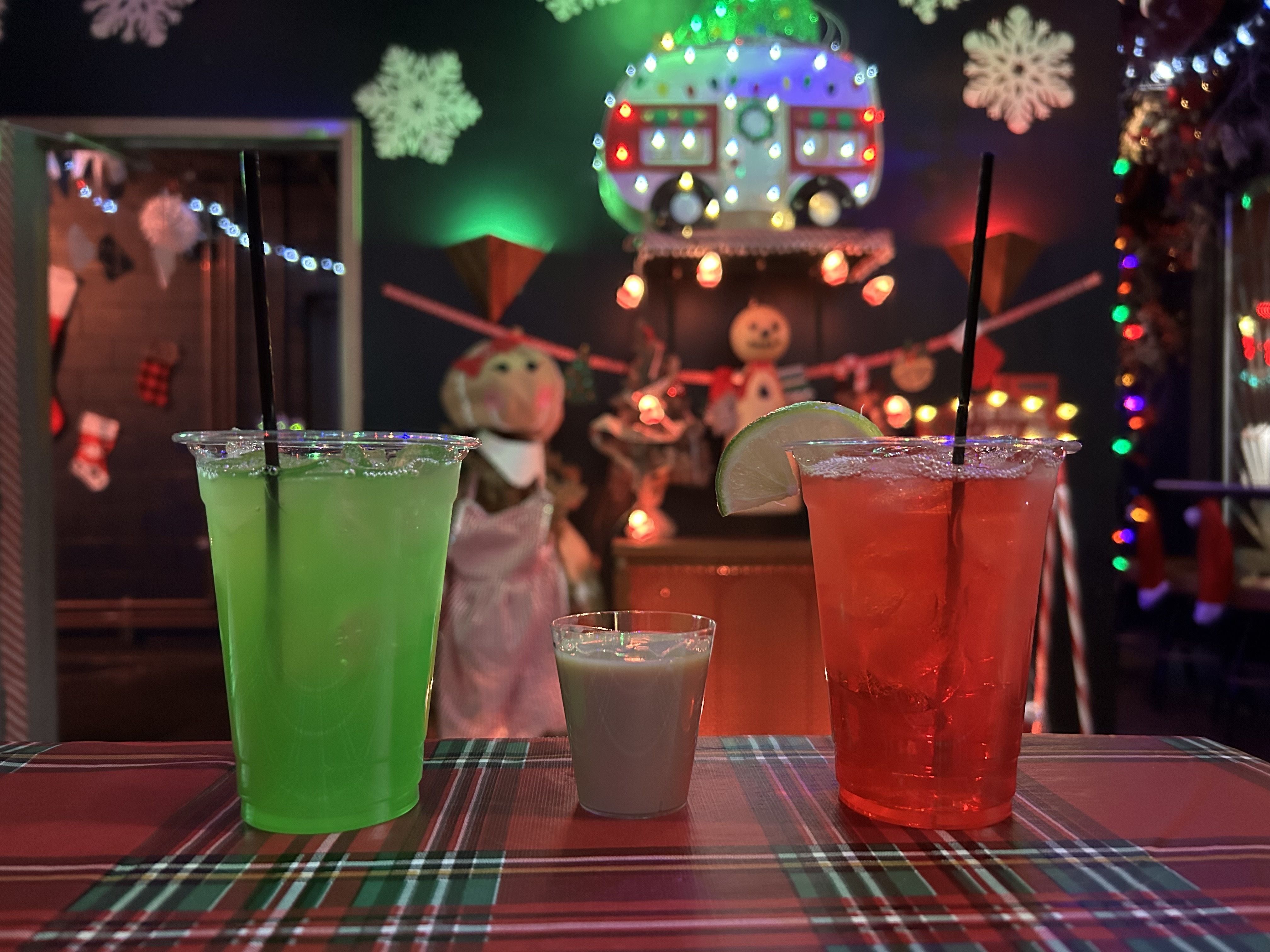 A green drink, a red drink and a milky-colored shot sit in front of Christmas decor including lights, snowmen and stockings.
