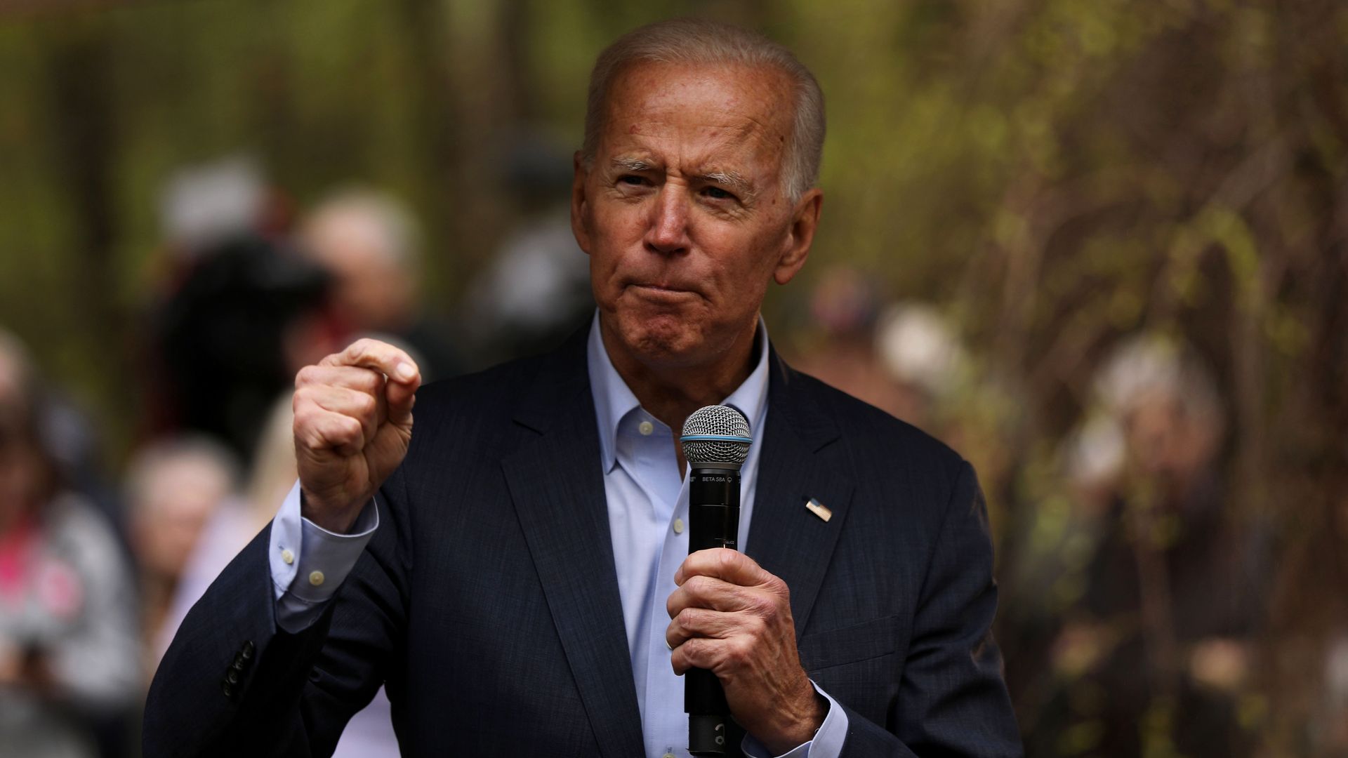 In this image, Biden speaks into a microphone while standing outside in a forested area.