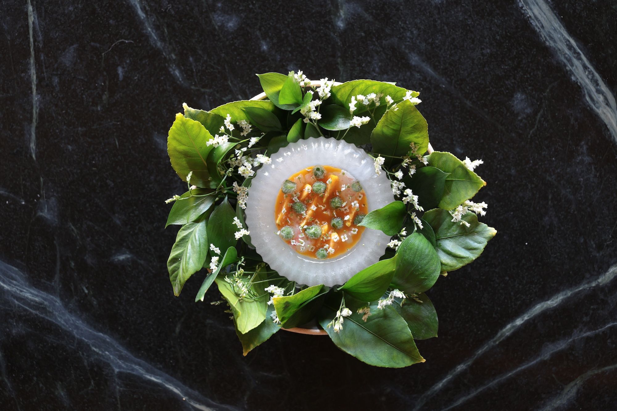 A bite-size dish with prawns, ice plant and flowers in a sauce that's served in a bowl made of ice surrounded by leaves. 