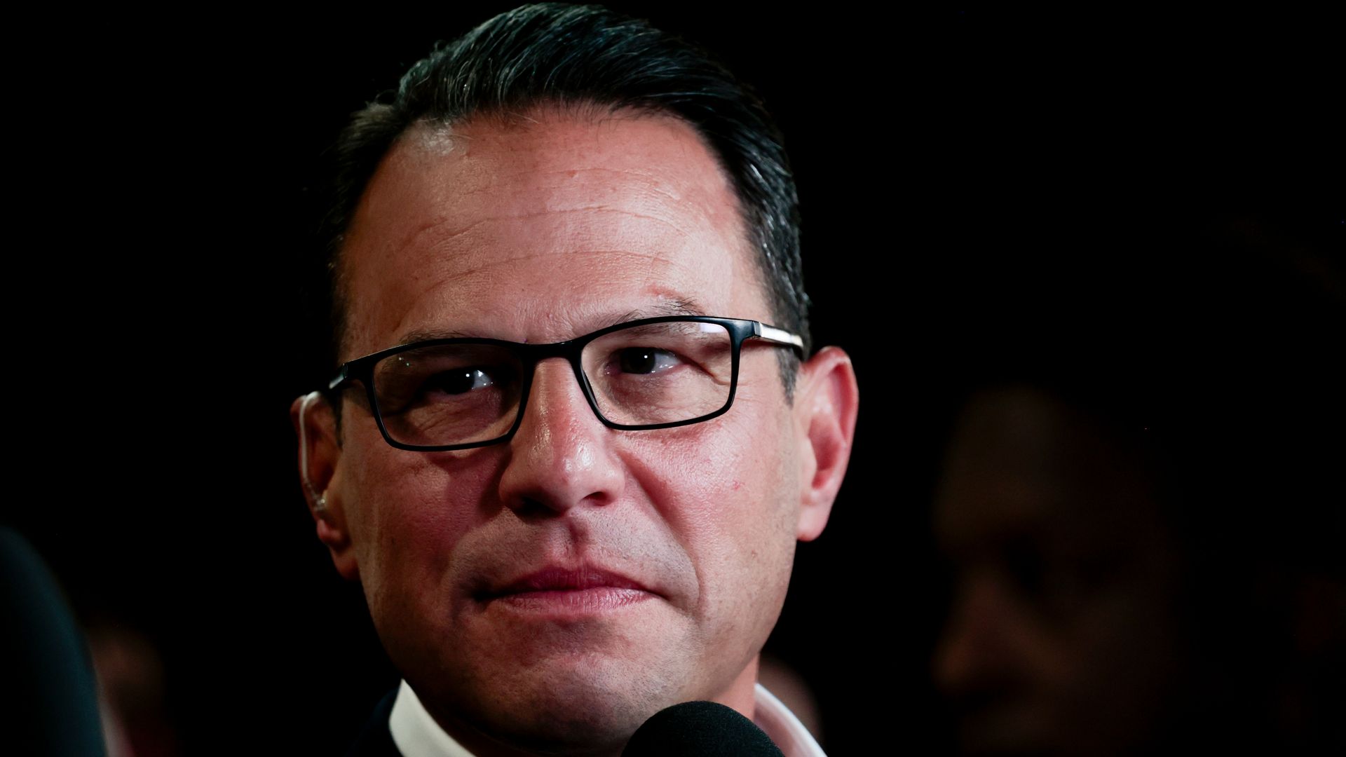 Close-up of a man with dark hair and black-framed glasses speaking into a microphone against a dark background; he wears a dark suit and white shirt.