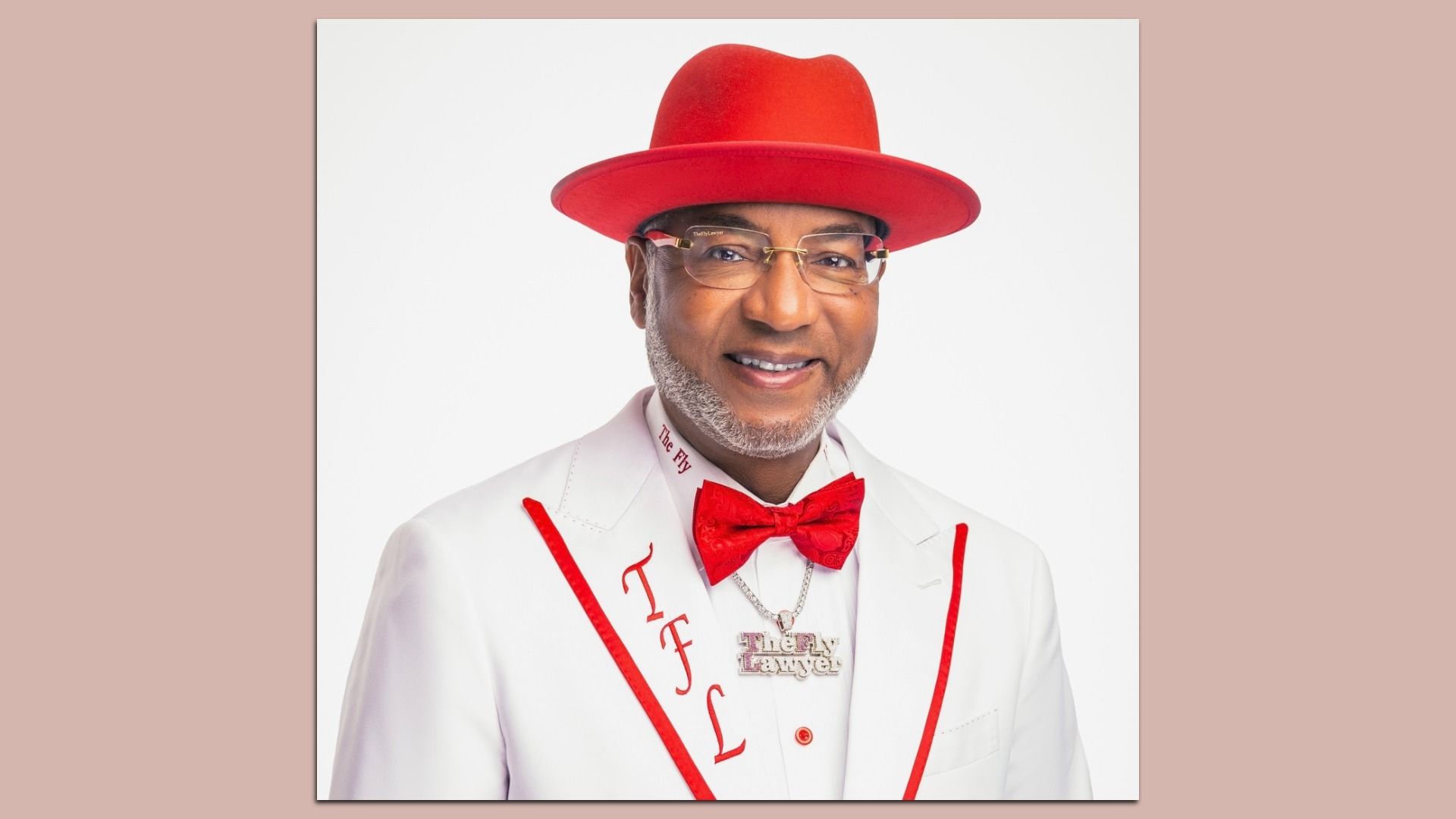 Smiling Black man in a white suit with red trim, red bow tie and red hat, gold-rimmed glasses, and a necklace reading "The Fly," against a white background.
