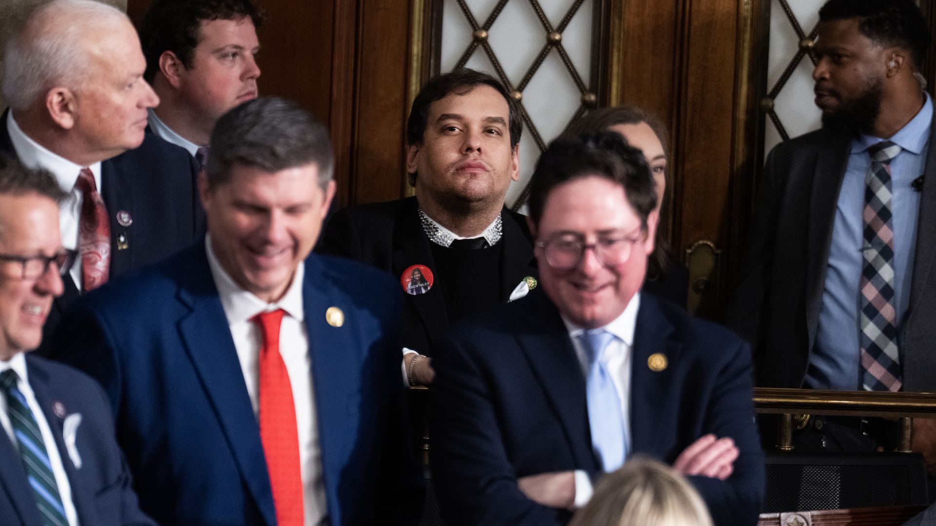 Former Rep. George Santos, wearing a black sweater over a white and gem-encrusted collared shirt, stands at the back of the House chamber.