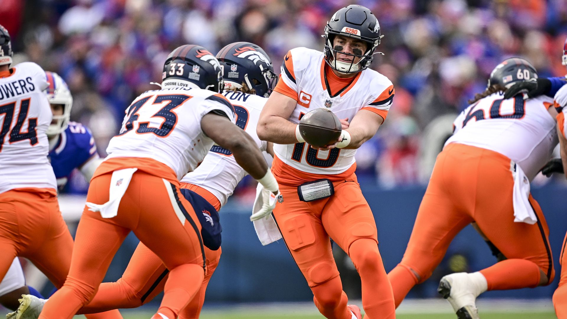 Denver Broncos football players in white and orange uniforms during a game, with the quarterback handing off the ball to a running back on the field.