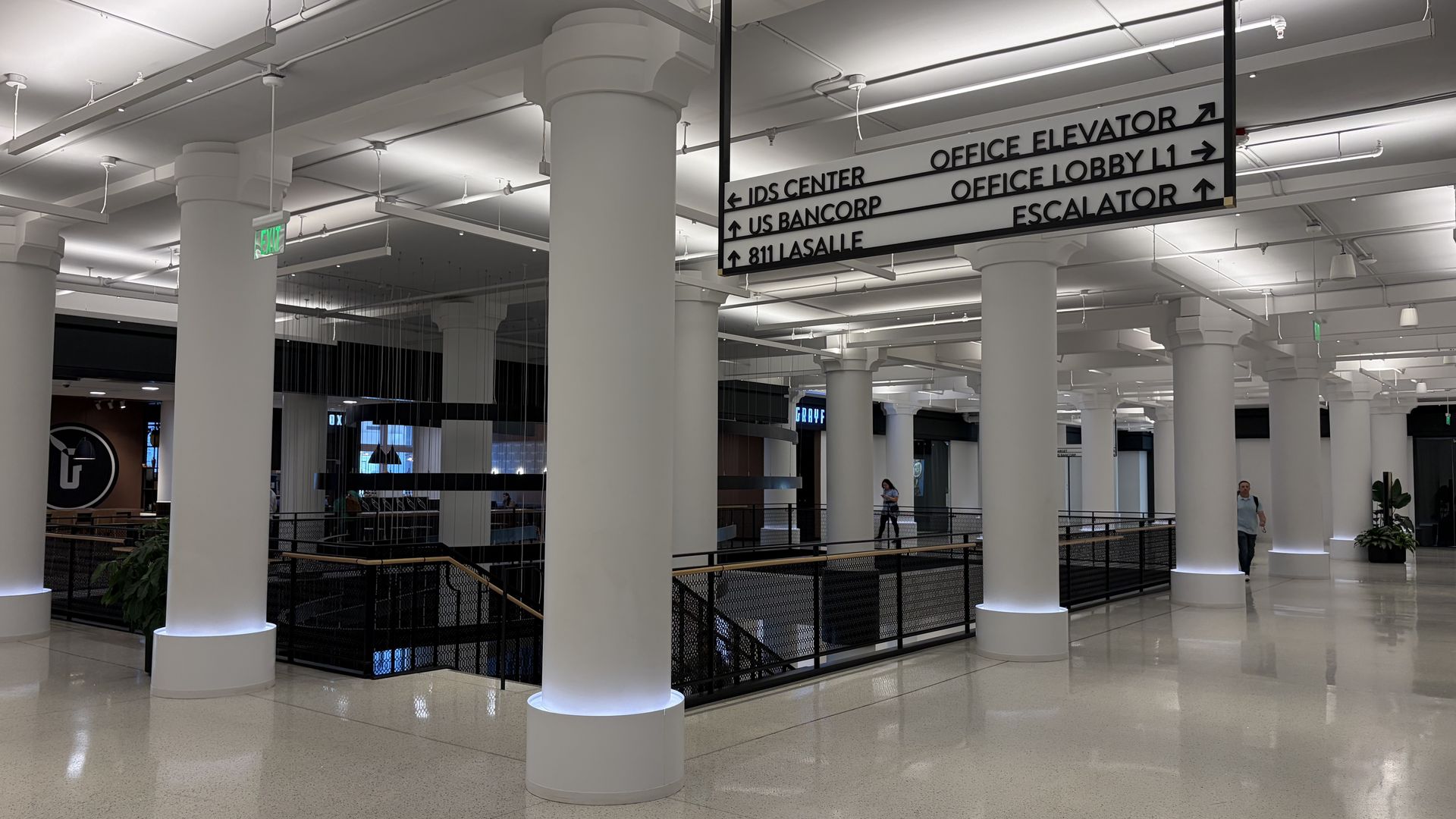 Indoor modern lobby with tall white pillars, bright overhead lighting, and a large directional sign listing offices and amenities (IDS Center, US Bancorp, 811 Lasalle, elevators, lobby, escalator).