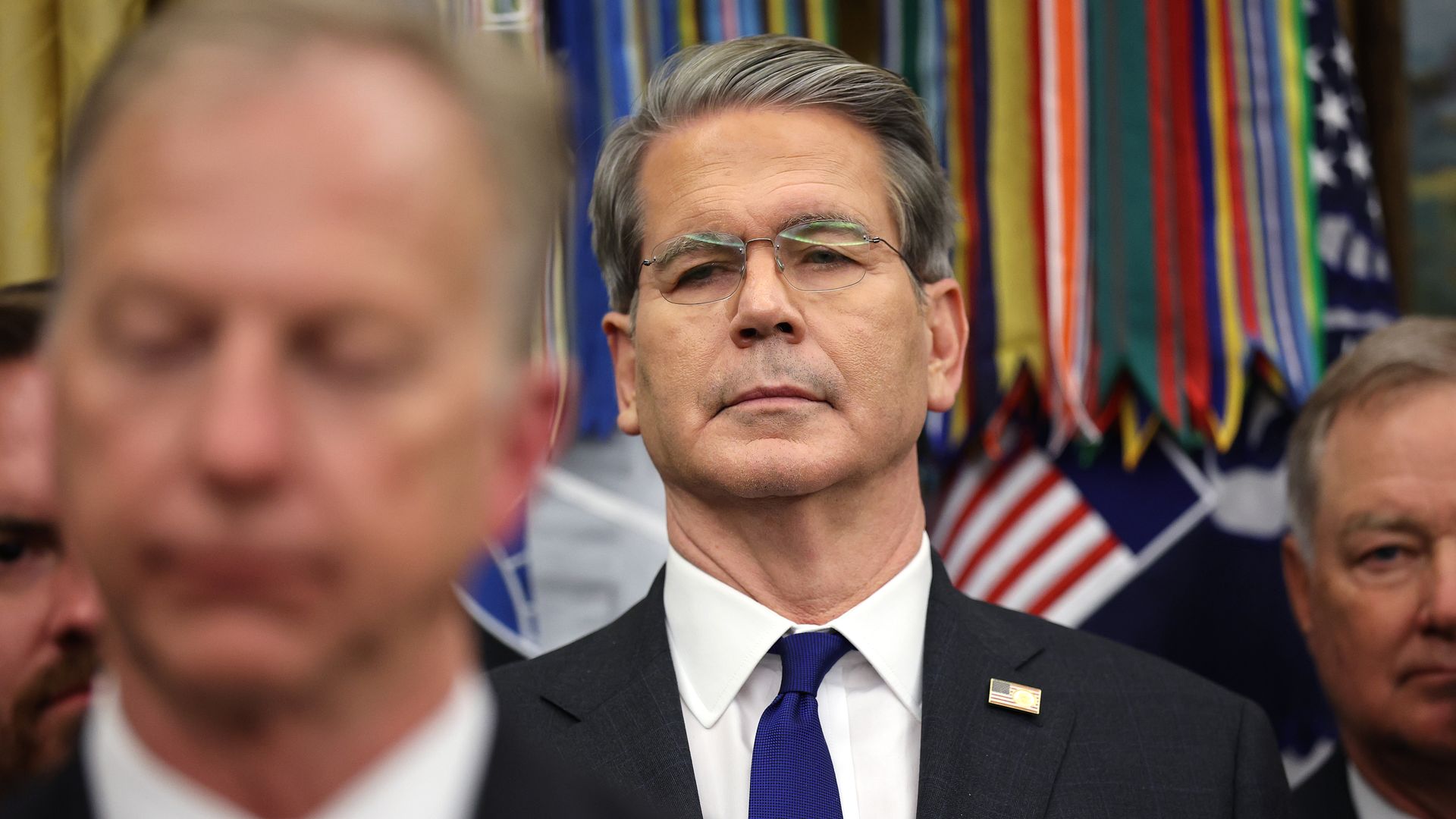 Treasury Secretary Scott Bessent, in a dark suit and dark blue tie with a U.S. flag lapel pin, standing amid other officials in the Oval Office. 