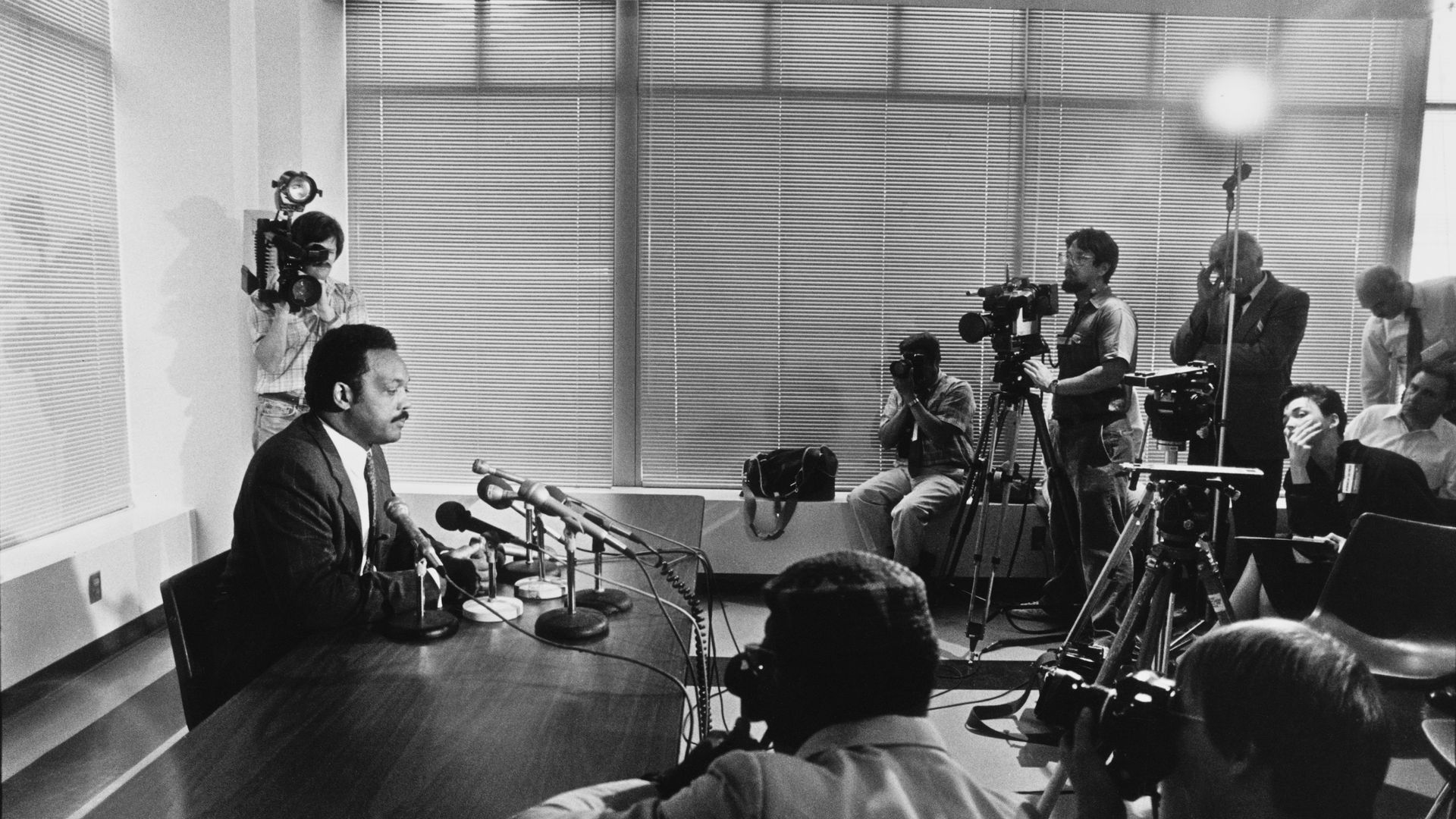 Black-and-white photo of Jesse Jackson in a suit speaking at a table with microphones, surrounded by photographers and cameramen in a room with closed blinds and bright lights.