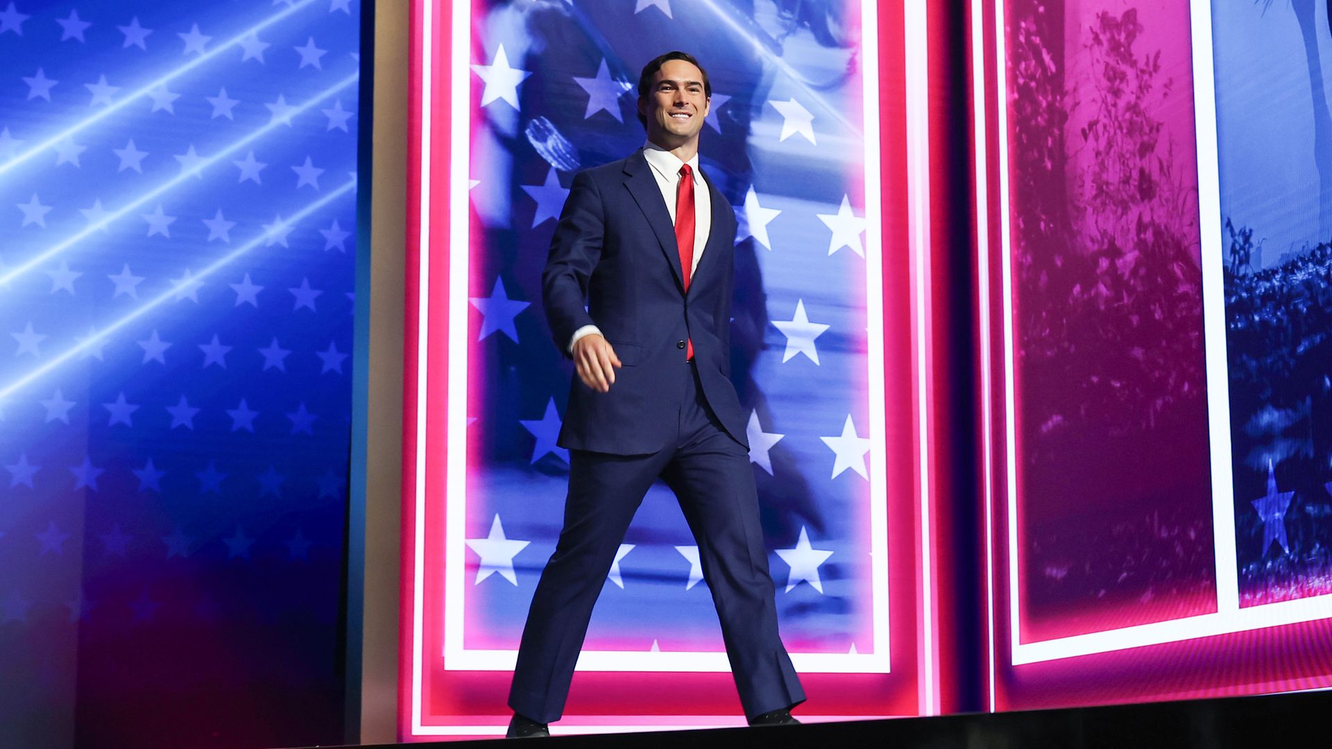 Smiling man in a navy suit and red tie walks onto a stage framed by blue starry panels and pink striped borders, with bright lights and a patriotic backdrop.