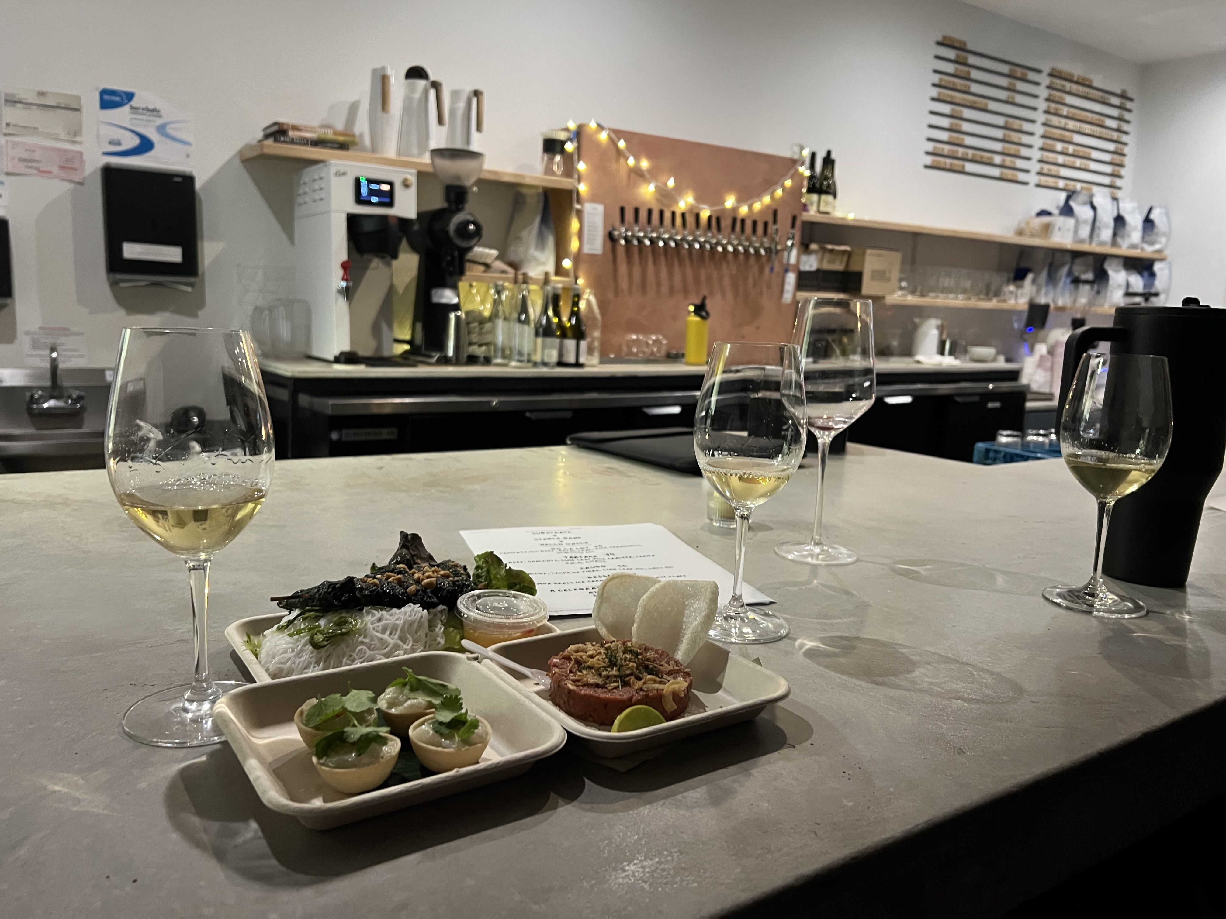 Counter with white wine glasses and plates of appetizers, a cozy bar background with string lights and a tap wall, bottles and coffee machine visible.