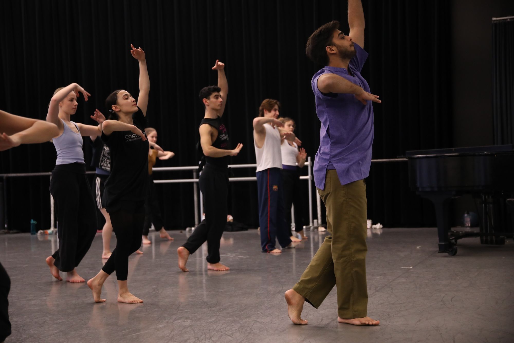 Group of dancers rehearsing in a studio with black curtains; a man in a purple shirt and khaki pants leads at right, raising one arm while others move in unison.