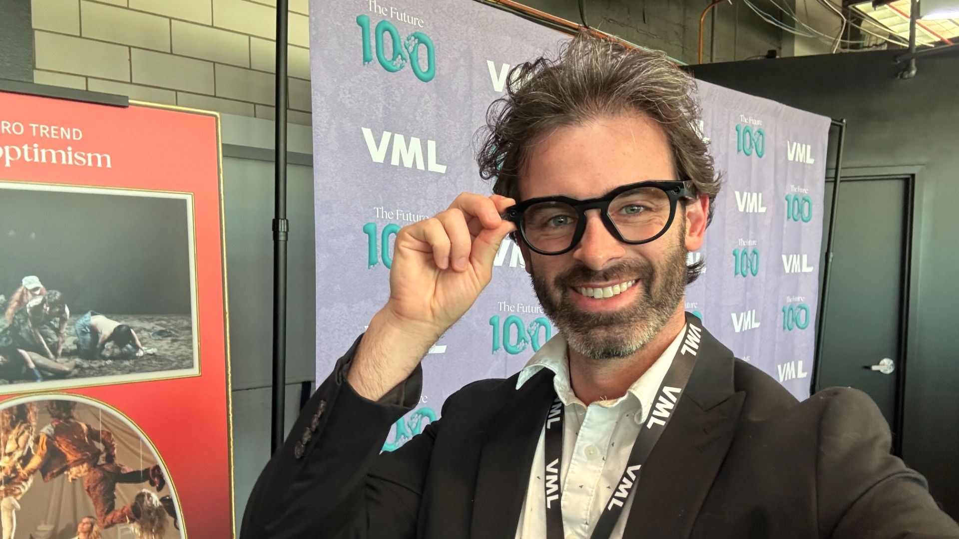 Reporter Travis wearing black-rimmed glasses in front of a backdrop that says "100" and "VML."