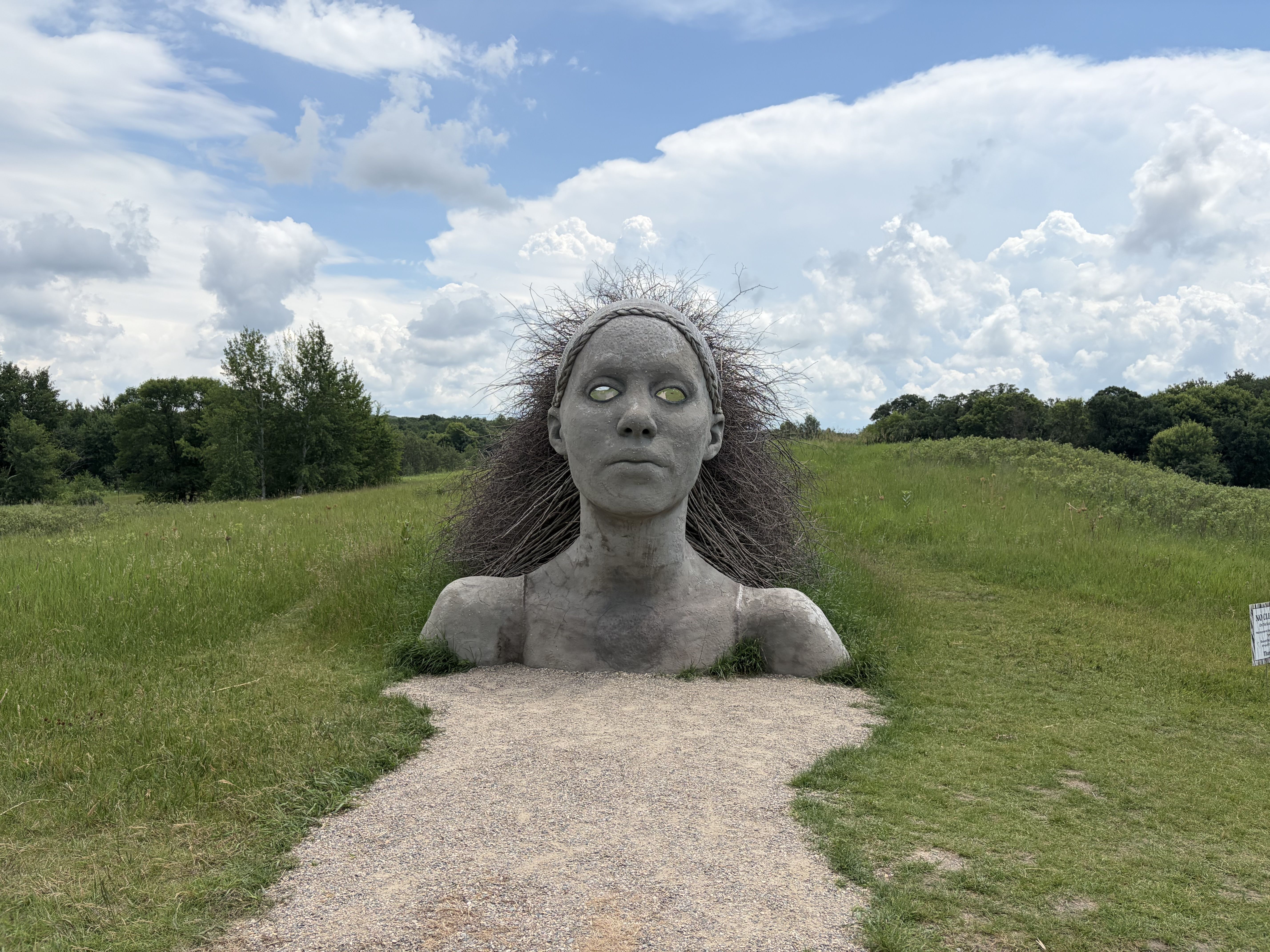 Life-size gray statue of a woman's head and shoulders emerging from the ground, rope around the crown and hair made of twigs, with hollow eyes, in a grassy field under a blue sky and clouds.
