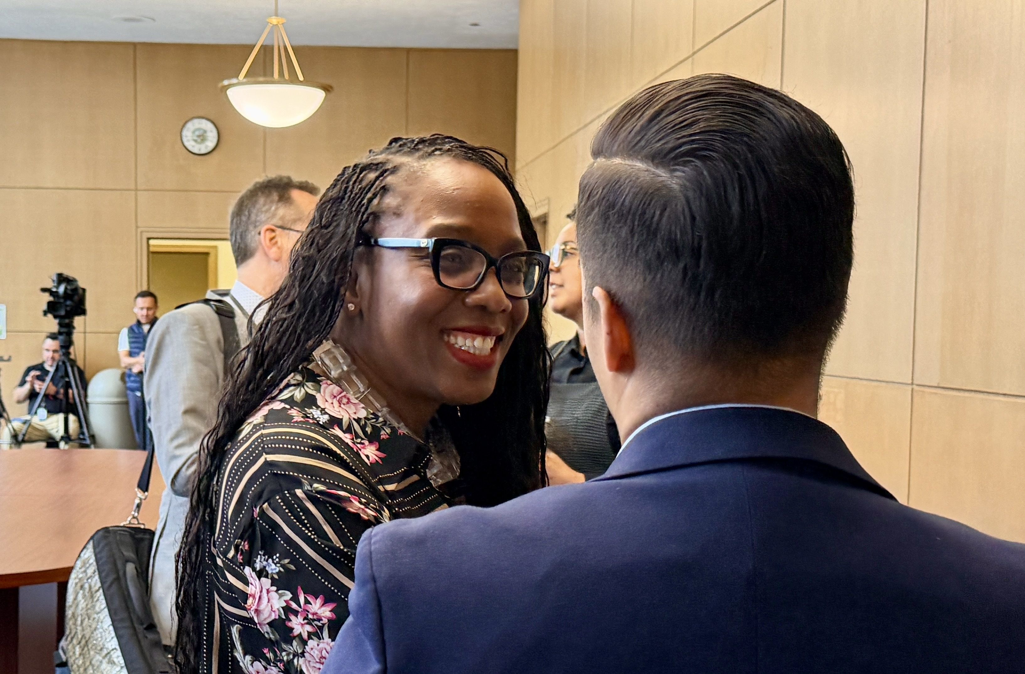 Smiling Black woman with glasses and braided hair, wearing a floral blouse, chats with a man in a navy suit in a wood-paneled room; photographers and a camera are in the background.