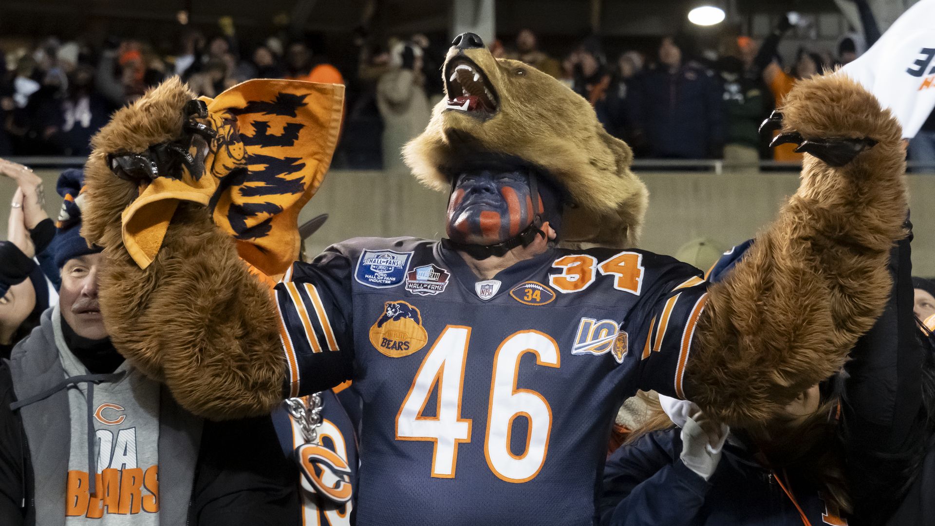 Chicago Bears fan wearing jersey 46 and bear head hat with furry arms, cheering with an orange towel in stadium crowd.
