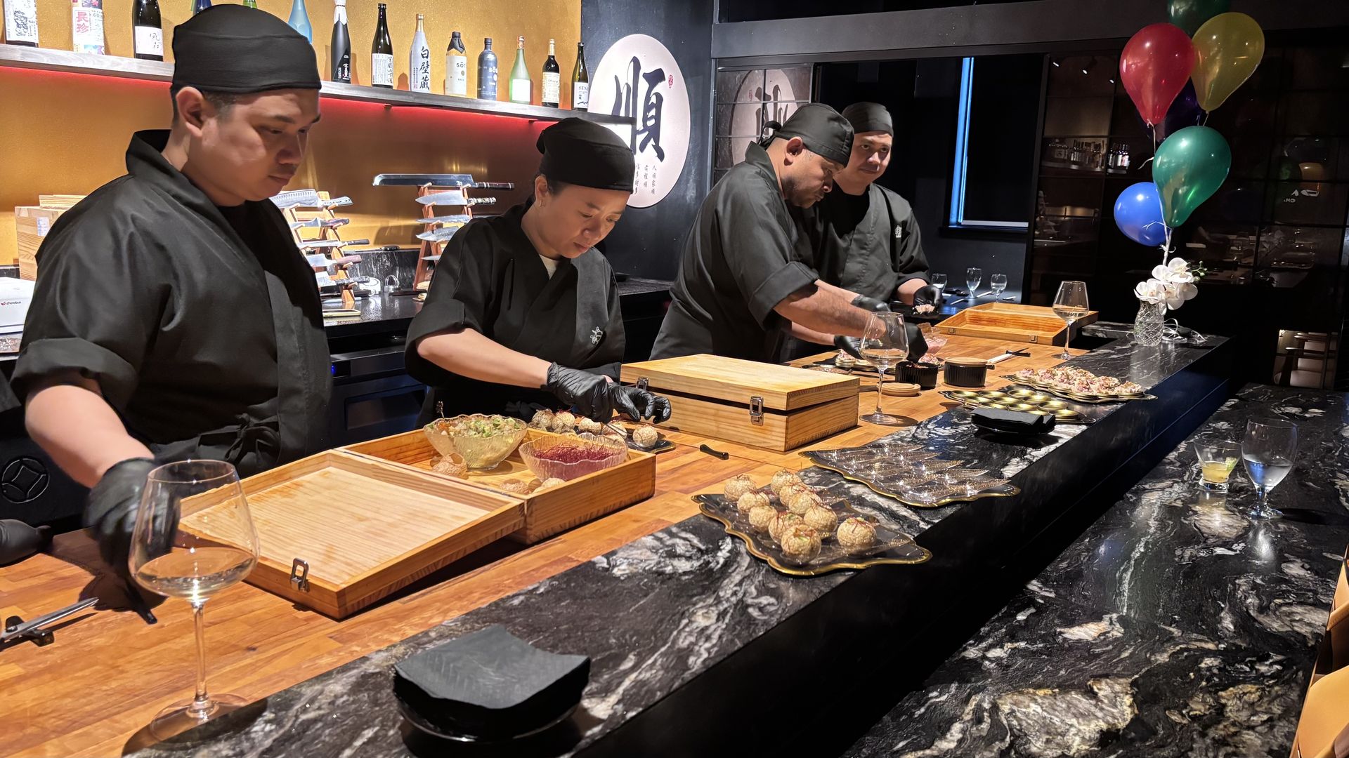 Four sushi chefs in black uniforms and caps prep dishes on wooden trays at a marble countertop; bottles line the back bar, and colorful balloons float beside a vase.