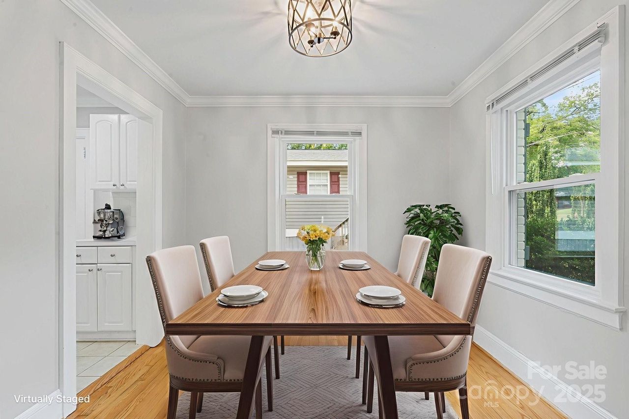 Bright dining room with wooden table, six beige upholstered chairs, white plates, a vase of yellow flowers, white walls, large windows, and a modern metal chandelier.