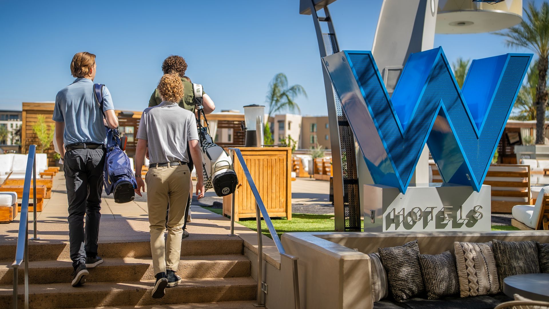 Three people carry golf bags on a rooftop patio area. 