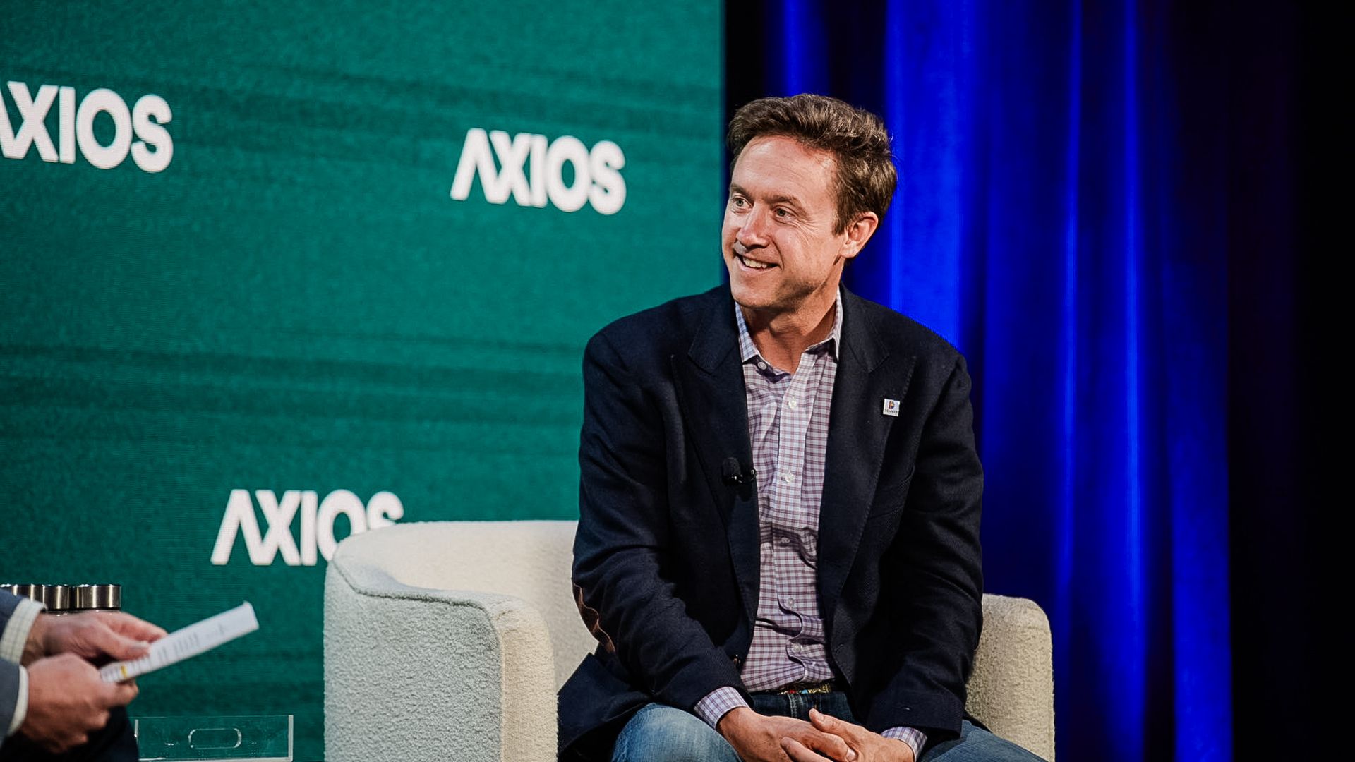 A man in a dark blazer and checkered shirt sits on a light gray chair on stage, smiling, with a green Axios backdrop and blue curtains to the side.
