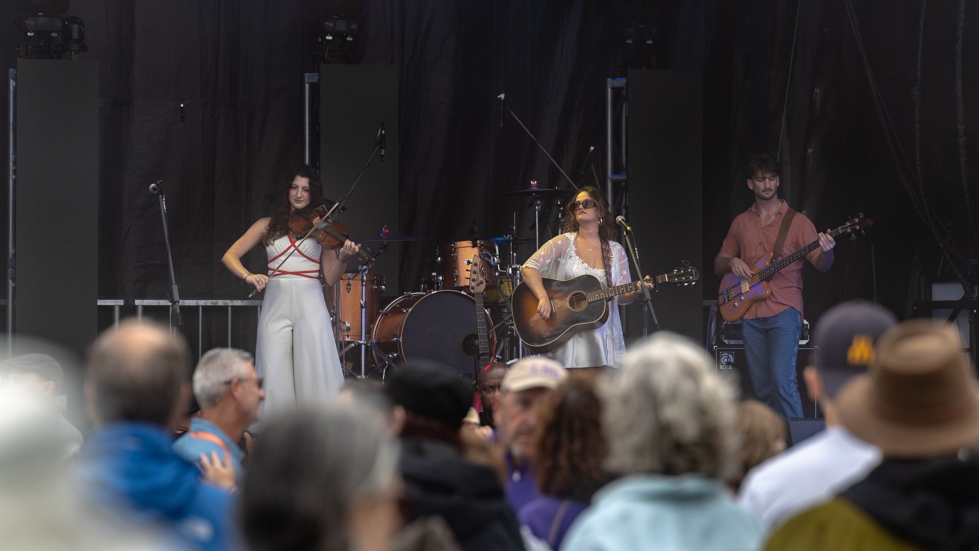 Three people perform music onstage. They are seen through a crowd of people.