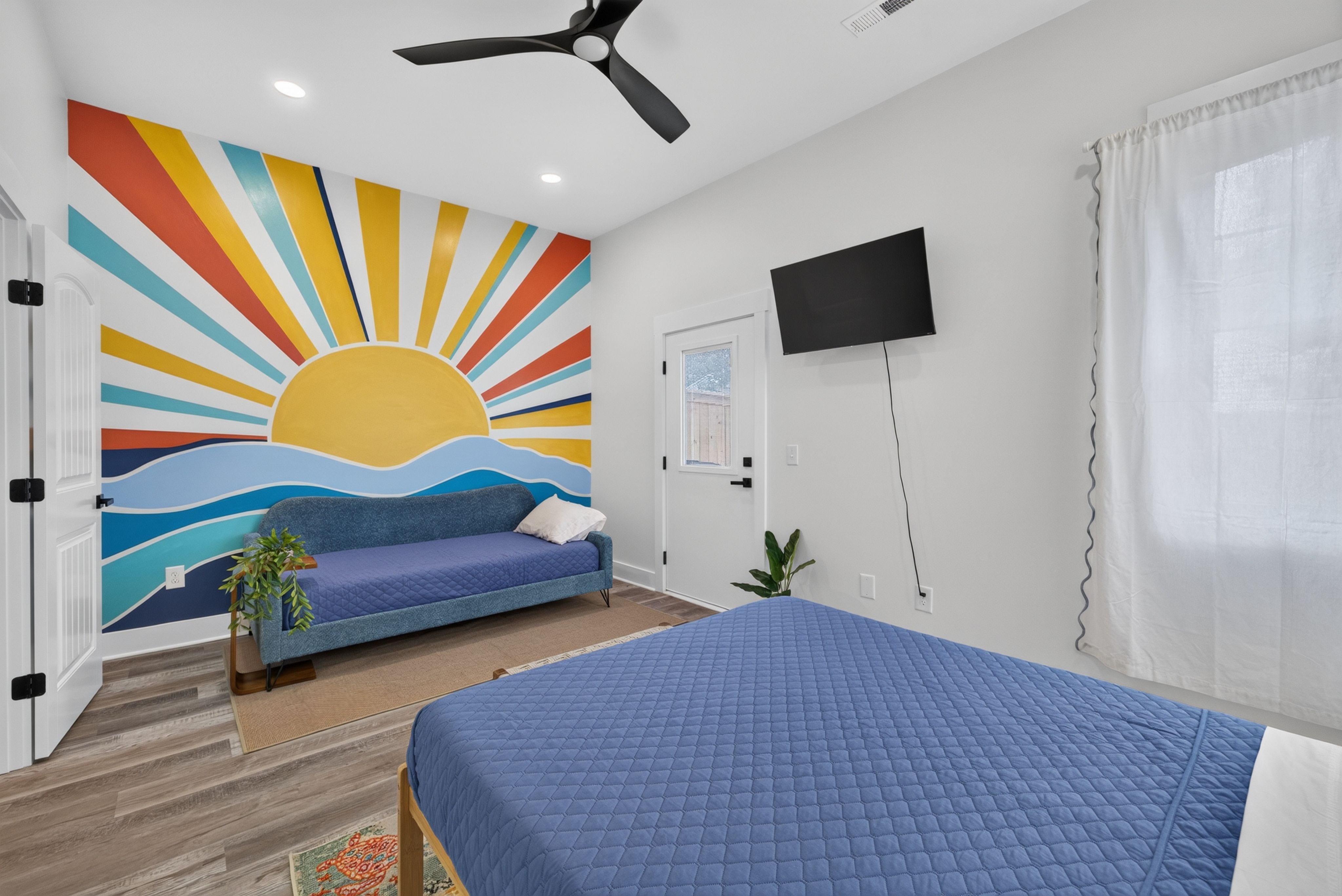 Bright bedroom with a sunburst mural on the left wall, blue sofa and two blue quilted beds. White door, wall-mounted TV, ceiling fan, and a large window with sheer curtains.