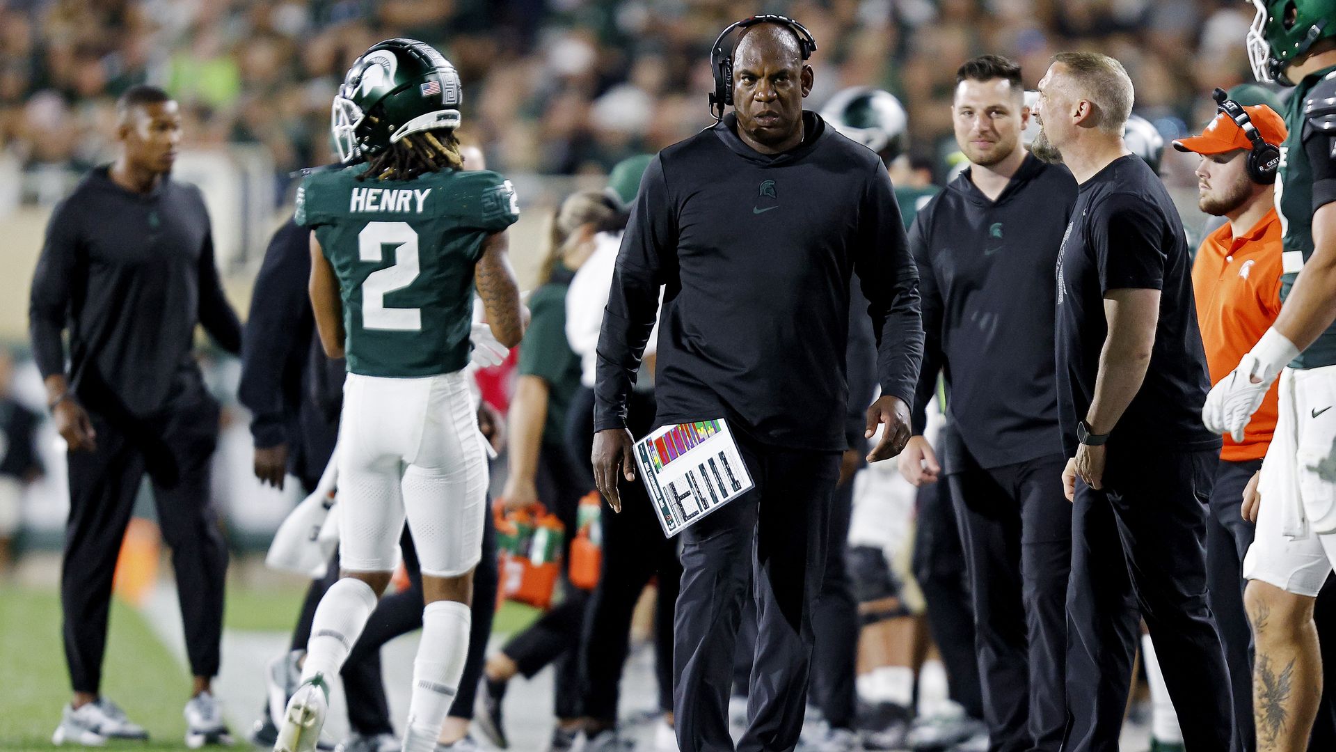 Mel Tucker walks toward the camera on the side of the field past other staff and players during a game.