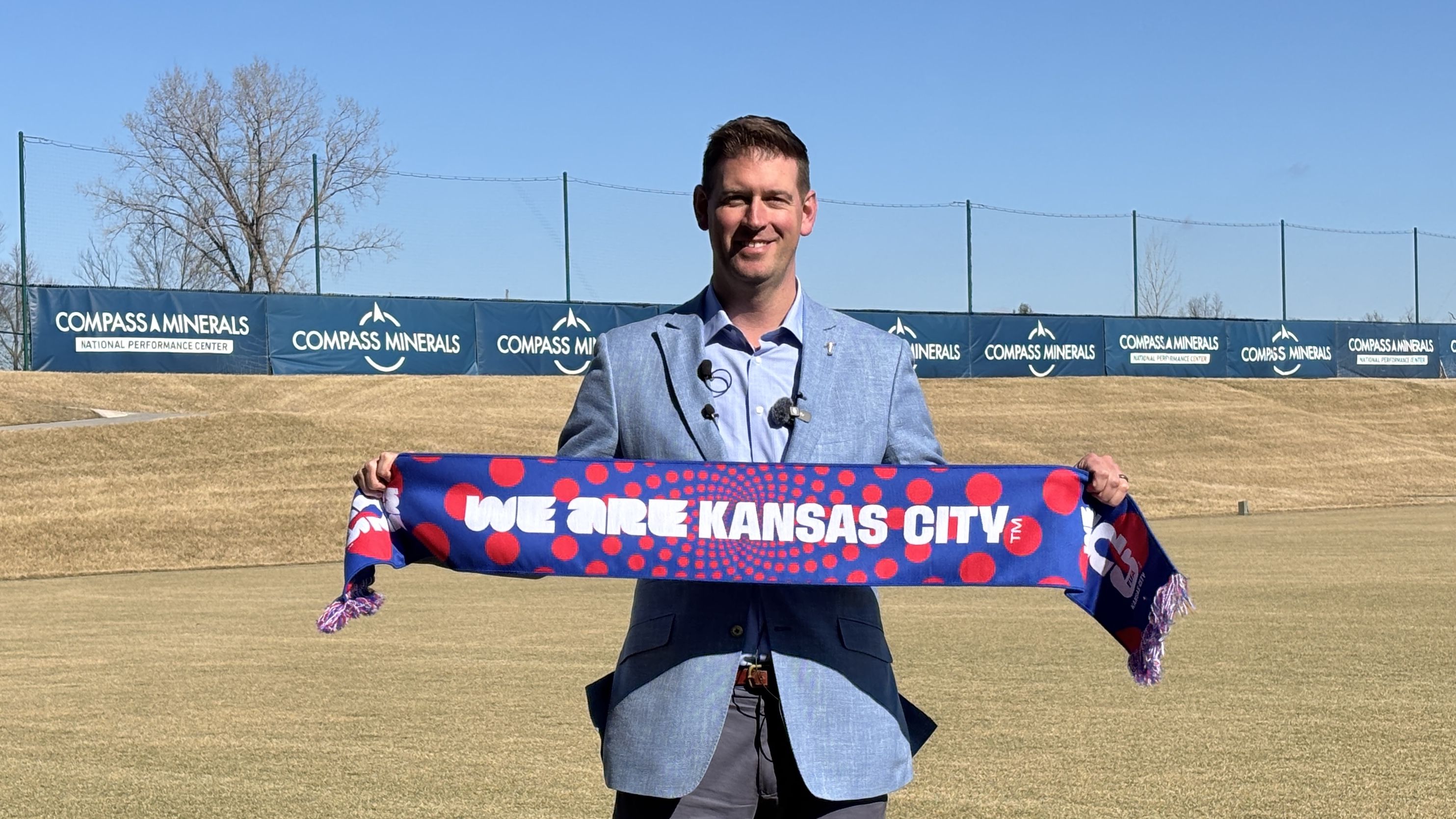 Sporting KC president and CEO Jake Reid in light blue blazer holding a blue and red scarf reading "WE ARE KANSAS CITY" on a sunny day at a field with Compass Minerals banners in the background.