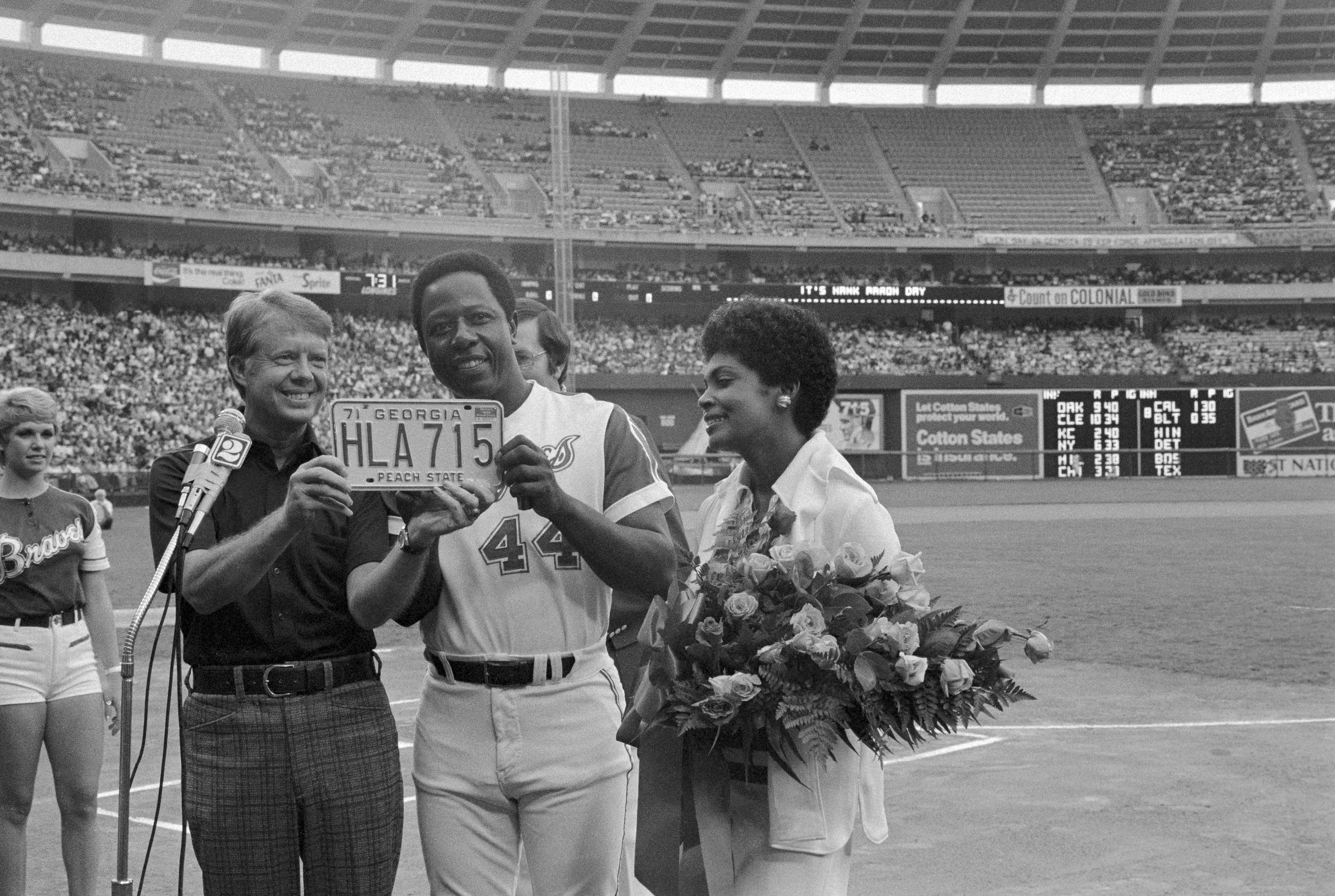 Black and white photo of Jimmy Carter next to Hank Aaron and Aaron's wife Billye (who's holding flowers). Carter and Aaron have a license plate that reads HLA715