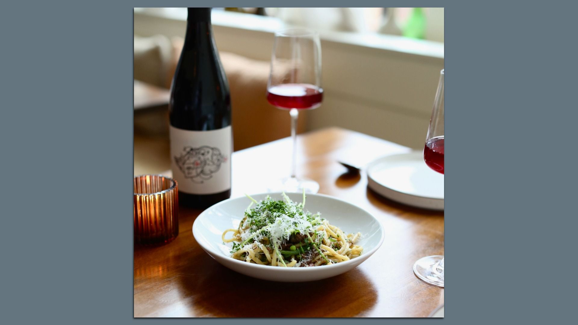 Plate of pasta topped with grated cheese and green herbs on a wooden table, accompanied by a bottle of red wine and two glasses partially filled with red wine.