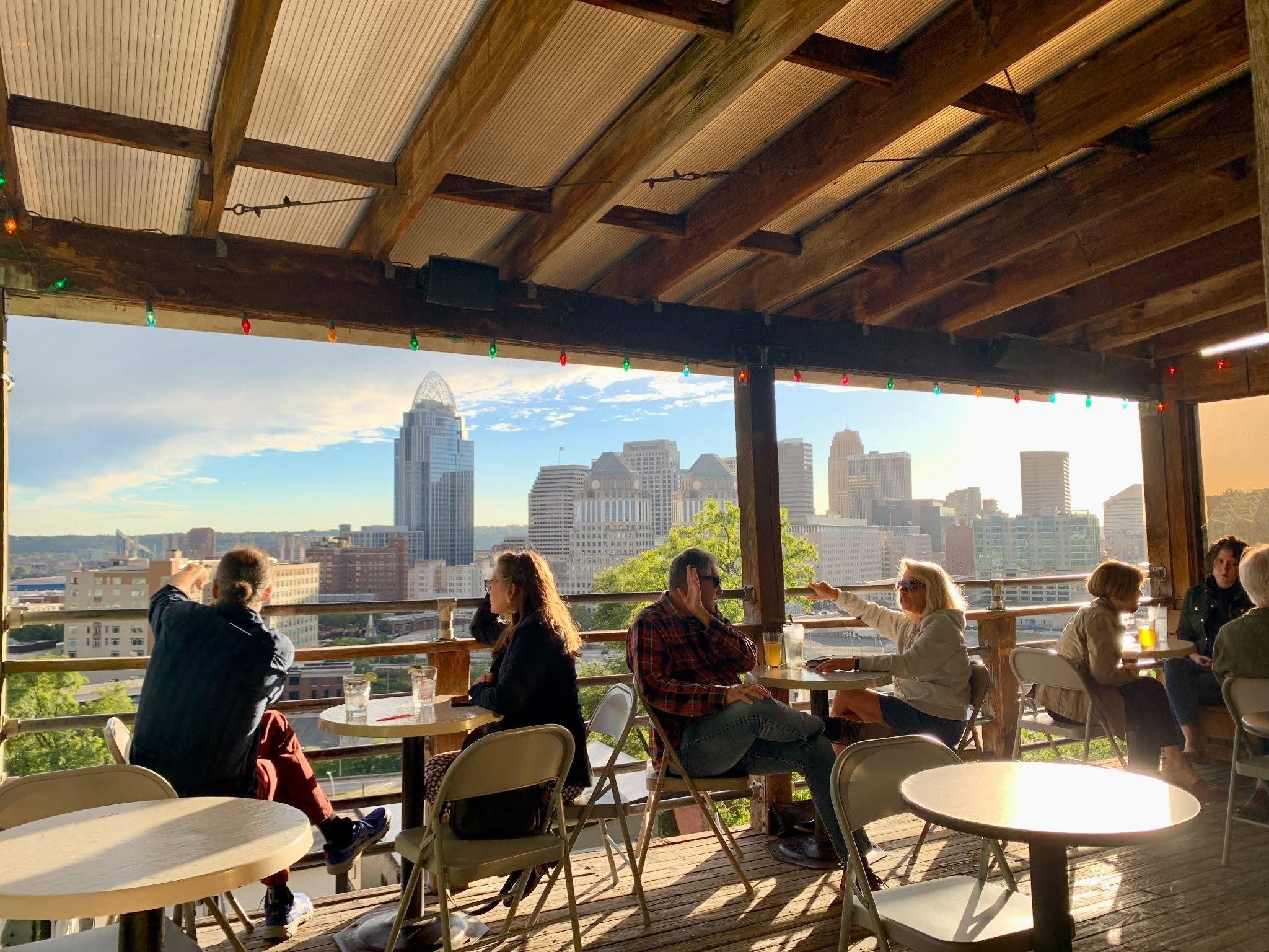 Open rooftop terrace with exposed wooden beams and colorful string lights. People sit at round tables enjoying drinks as a city skyline gleams in the background on a sunny day.
