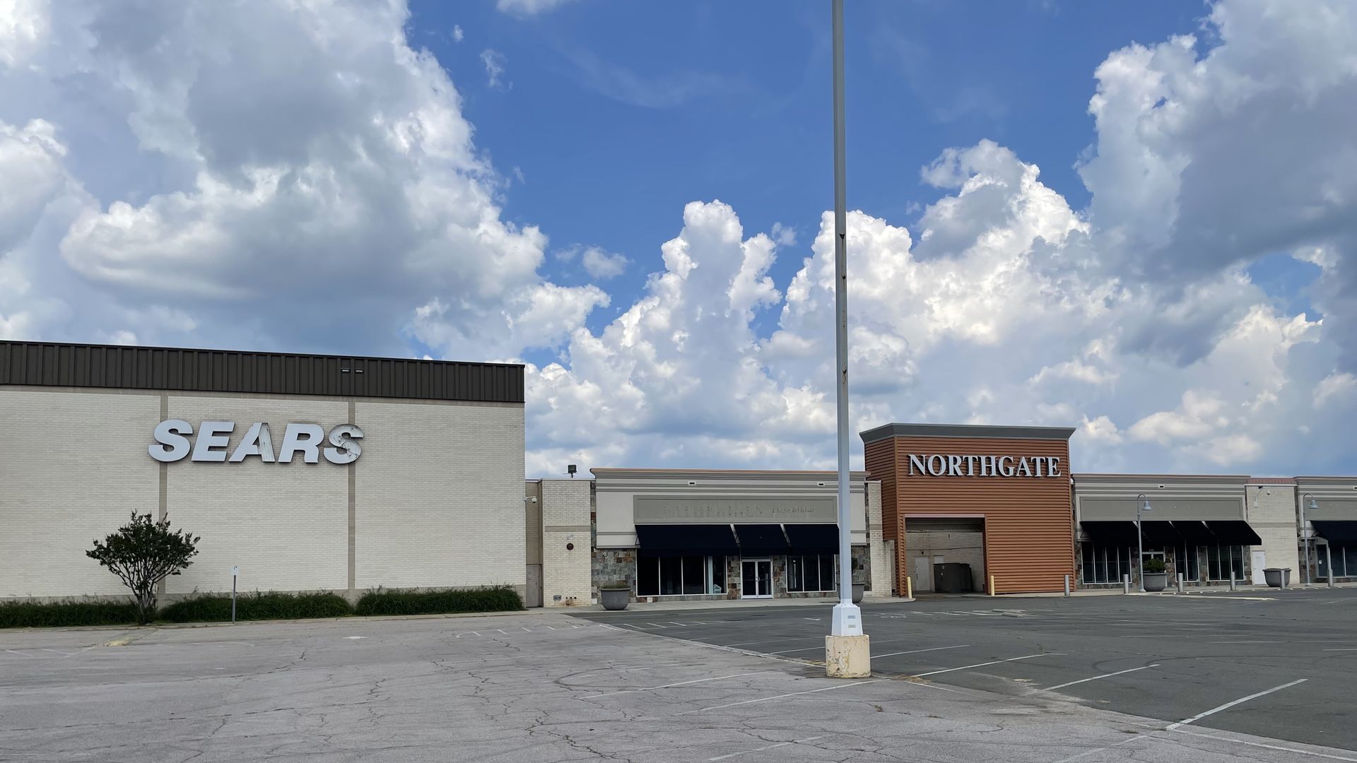 Empty parking lot in front of a beige shopping center with a worn SEARS sign and a NORTHGATE entrance under a partly cloudy blue sky.