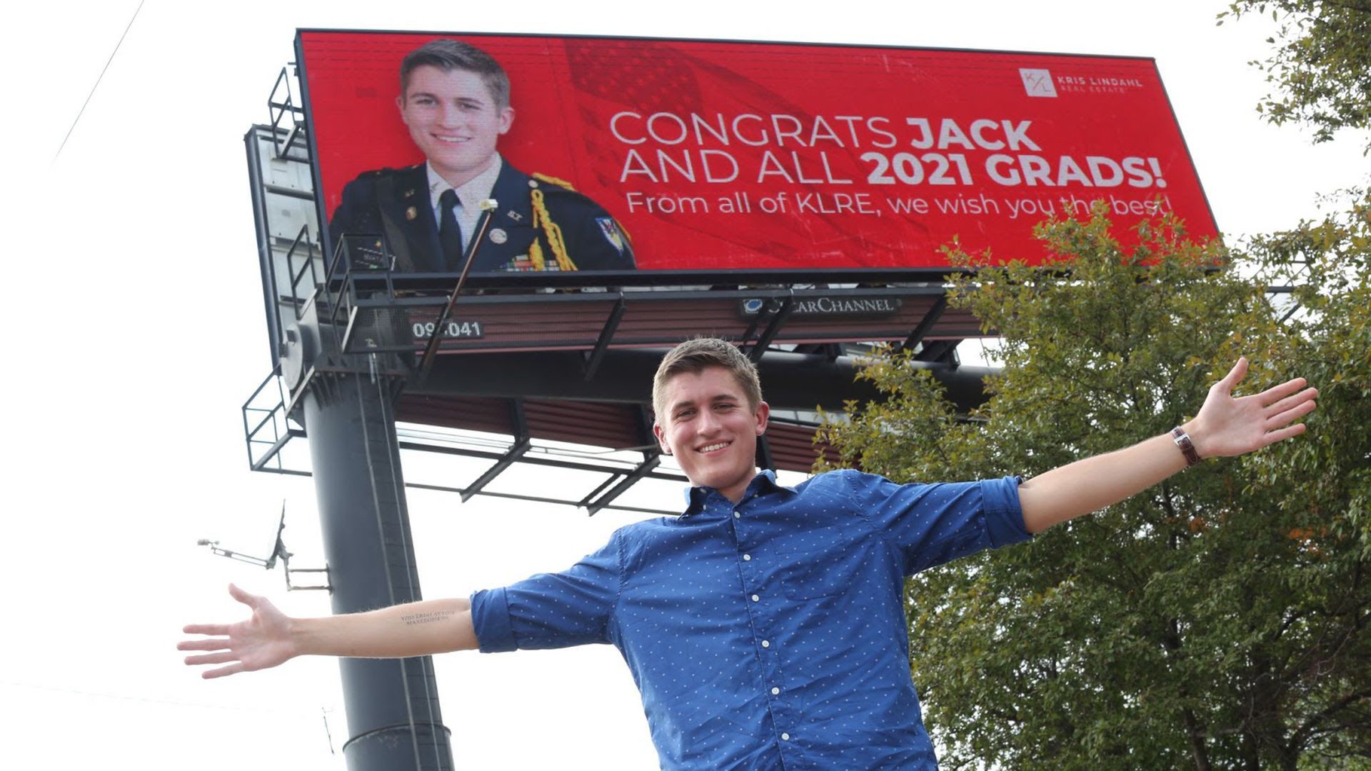 A graduating high school senior stands with his arms outstretched by a billboard celebrating his graduation set up by Kris Lindahl's real estate group.