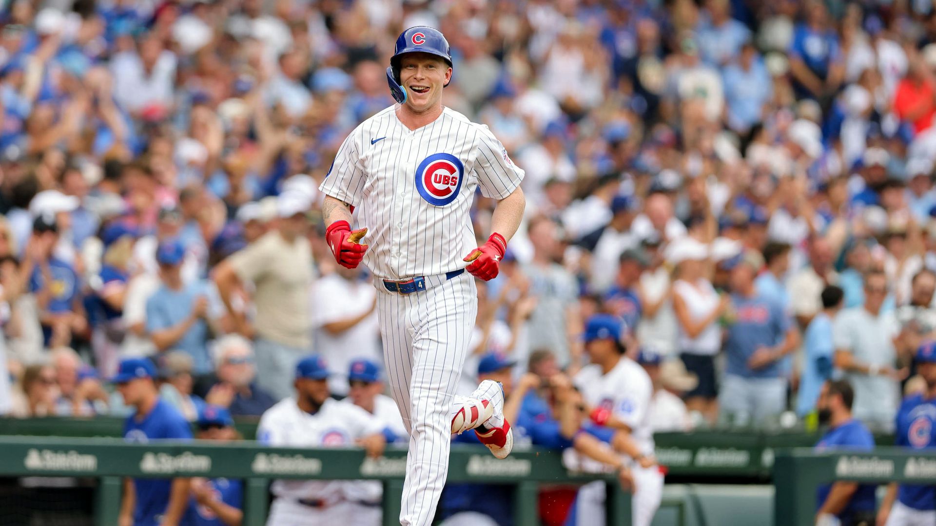 Chicago Cubs baseball player in a white pinstripe uniform runs toward the camera, wearing a blue Cubs helmet and red gloves, as a cheering crowd blurs in the background.