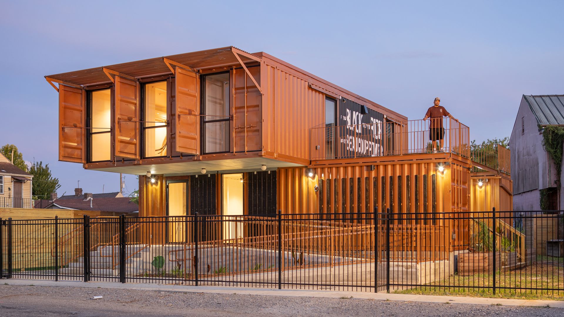Container home made from orange shipping containers with glass doors and a second-floor balcony; warm interior lights glow. A man stands on the balcony as a black fence and a small dog sit out front.