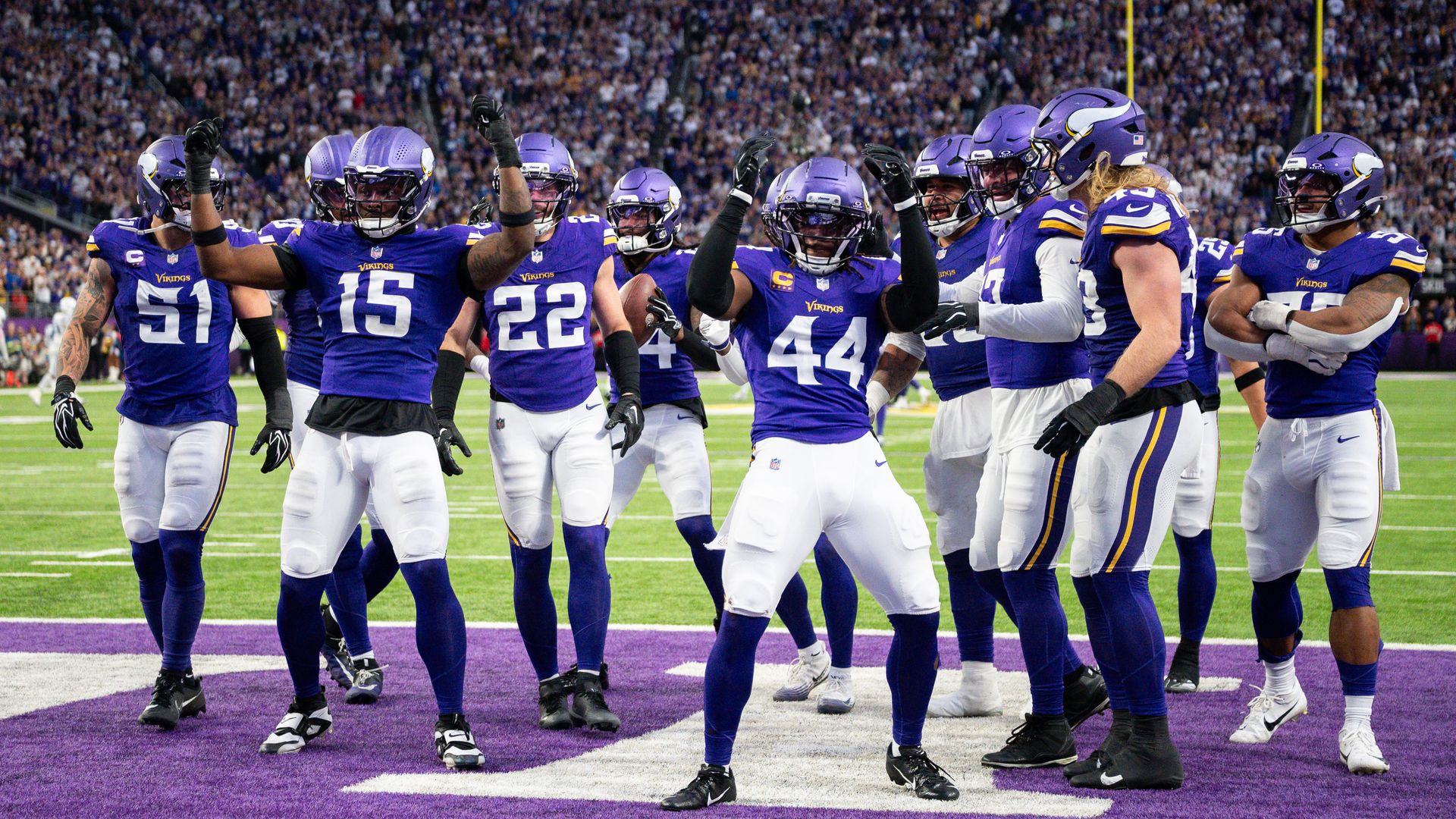 Group of Minnesota Vikings players in purple uniforms celebrate on the field, arms raised, as a large crowd cheers in the stands.
