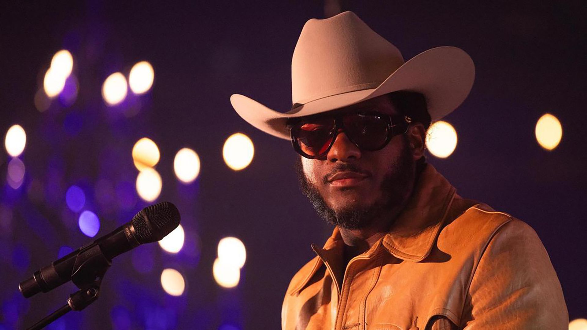 A man wearing a light cowboy hat, red-tinted sunglasses and an orange-brown jacket stands at a microphone on stage, with warm bokeh city lights in the dark background.