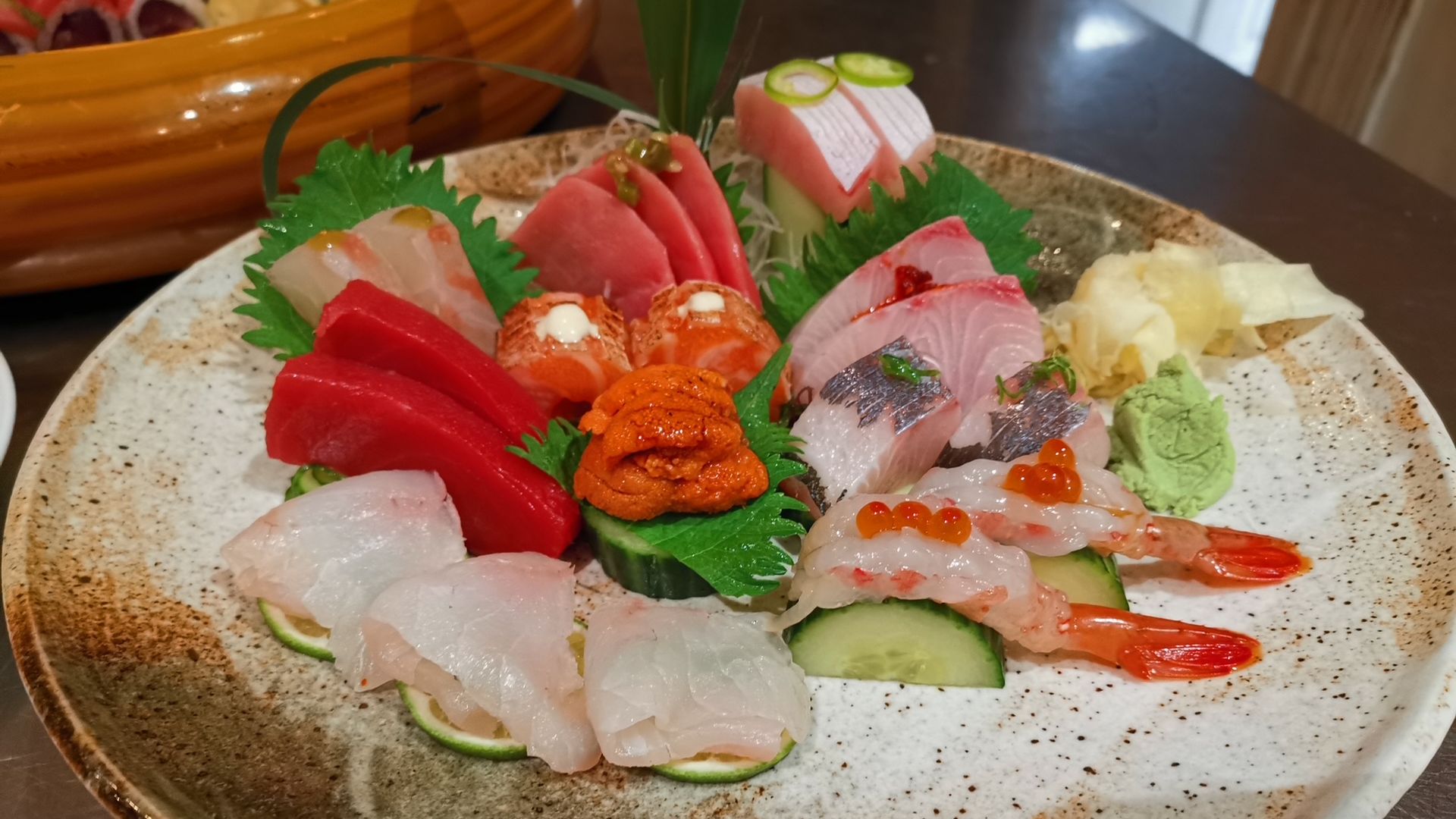 Assorted sashimi on a ceramic plate: red tuna, orange sea urchin, pink salmon, white fish, and shrimp with orange roe; garnished with cucumber slices, green shiso leaves, wasabi, and pickled ginger.