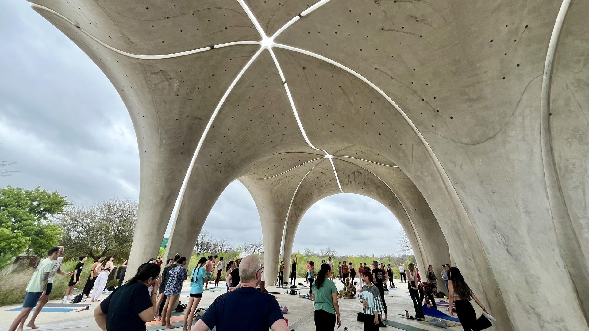 A group of people standing above their yoga mats underneath a large concrete pavilion with a cloudy sky in the background.
