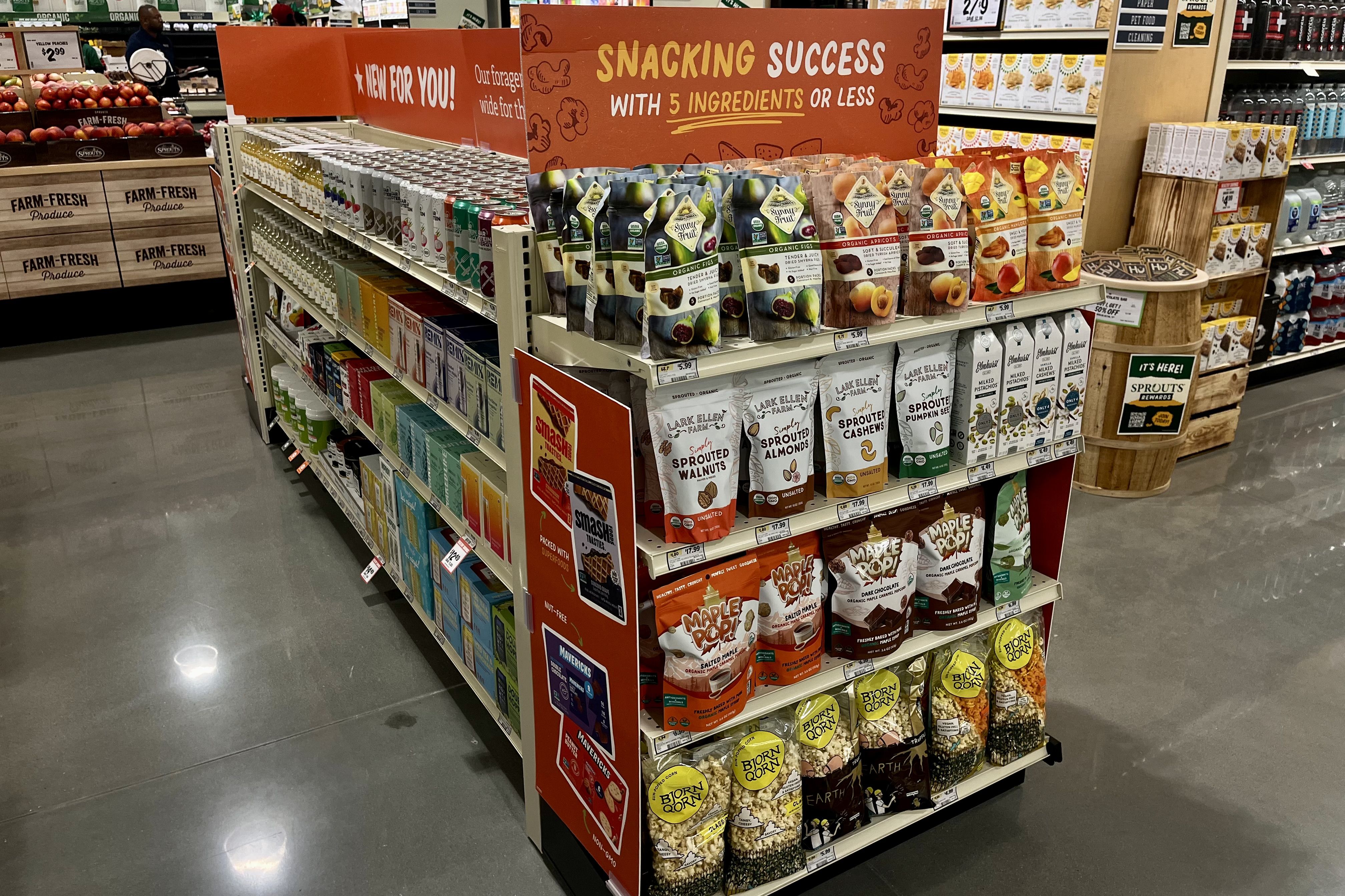 Grocery store snack aisle display with organic dried fruit, sprouted nuts, popcorn, and beverages.