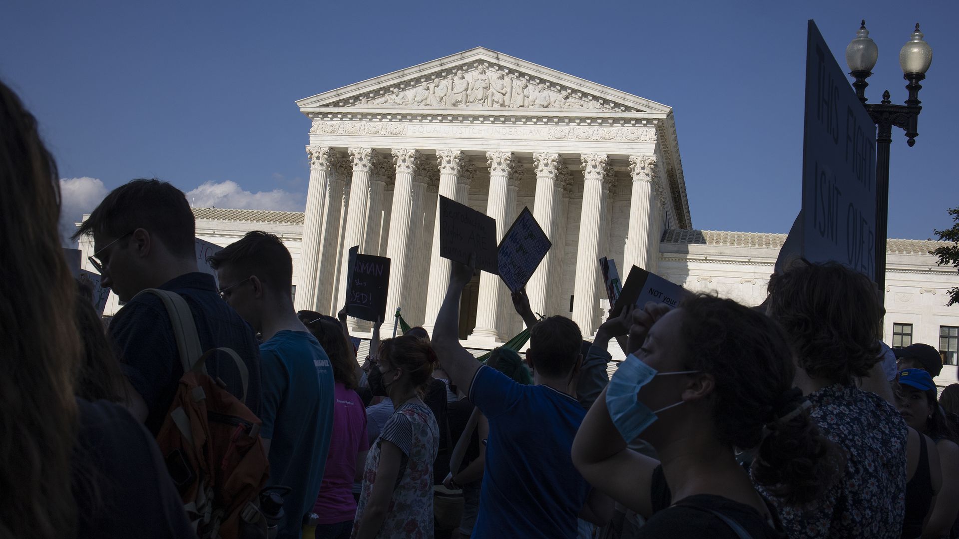 Protesters covered in shadows are gathered at the Supreme Court building, which is illuminated by the sun under a clear blue sky.