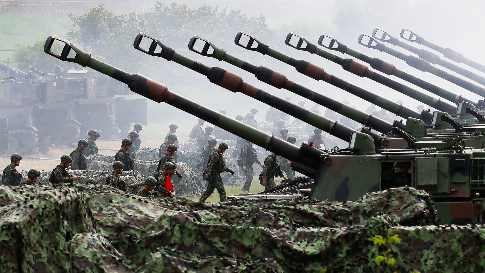 Rows of camouflaged artillery guns with long barrels pointed upward, and soldiers in green uniforms and helmets walking among them in a foggy outdoor setting.